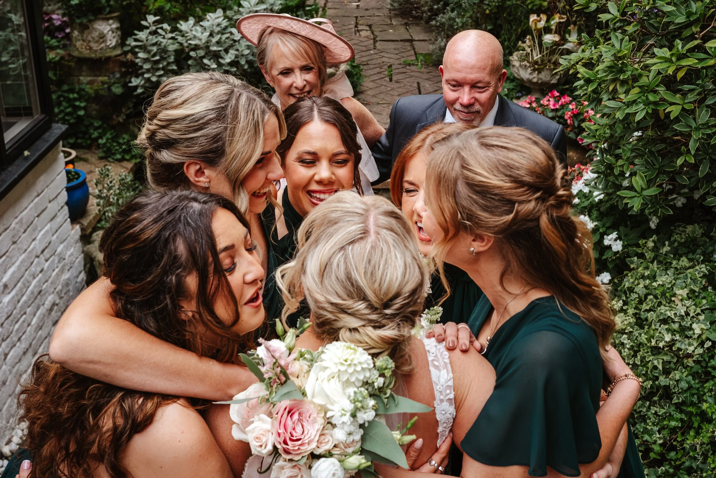 A group of women and an older man are hugging and smiling in a garden, celebrating a wedding with the bride holding a bouquet of pink and white flowers. The Kings Chapel, Amersham