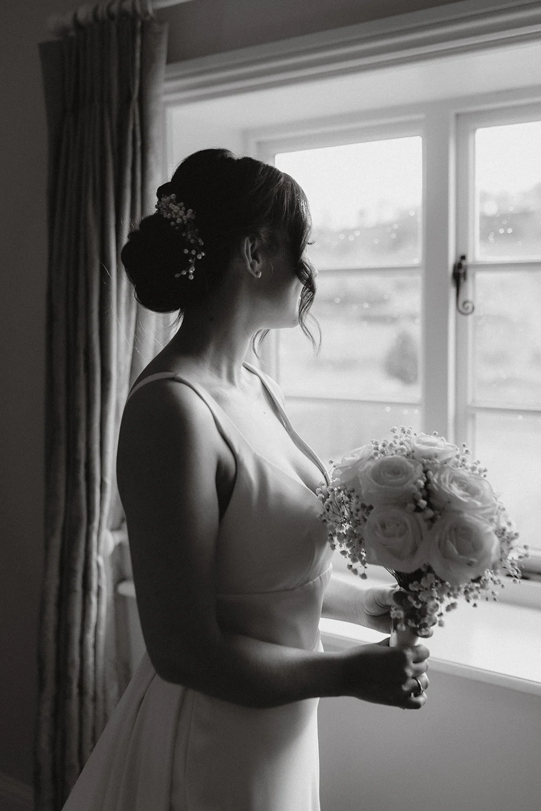 A black-and-white photo of a bride standing by a window, holding a bouquet of roses and baby's breath flowers, while looking outside.