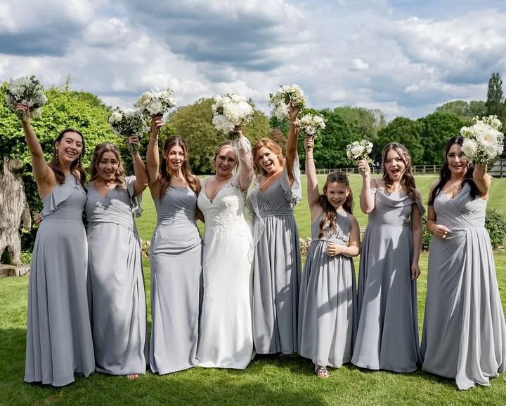 A group of seven women and one young girl standing outdoors on green grass, all dressed in light gray bridesmaid dresses with the bride in a white wedding gown. They are holding bouquets of white flowers and smiling, with their arms raised in celebra