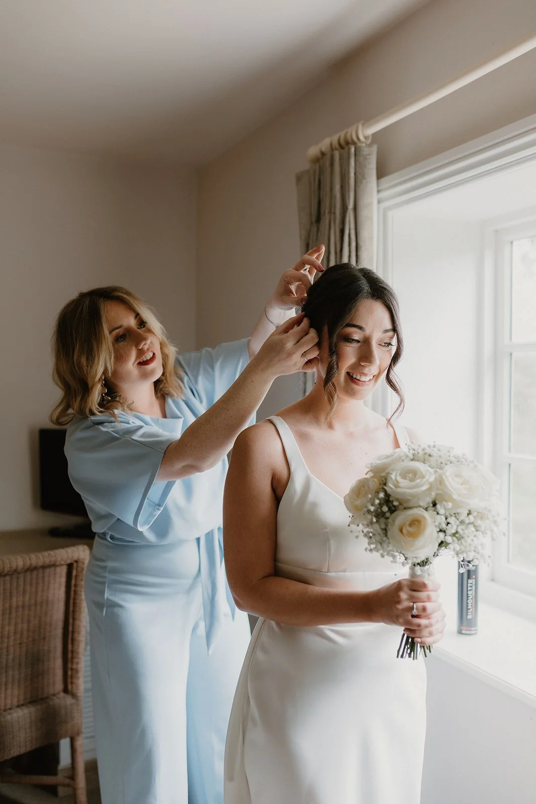A bride holding a bouquet of white roses, standing near a window, as a woman adjusts her hair.