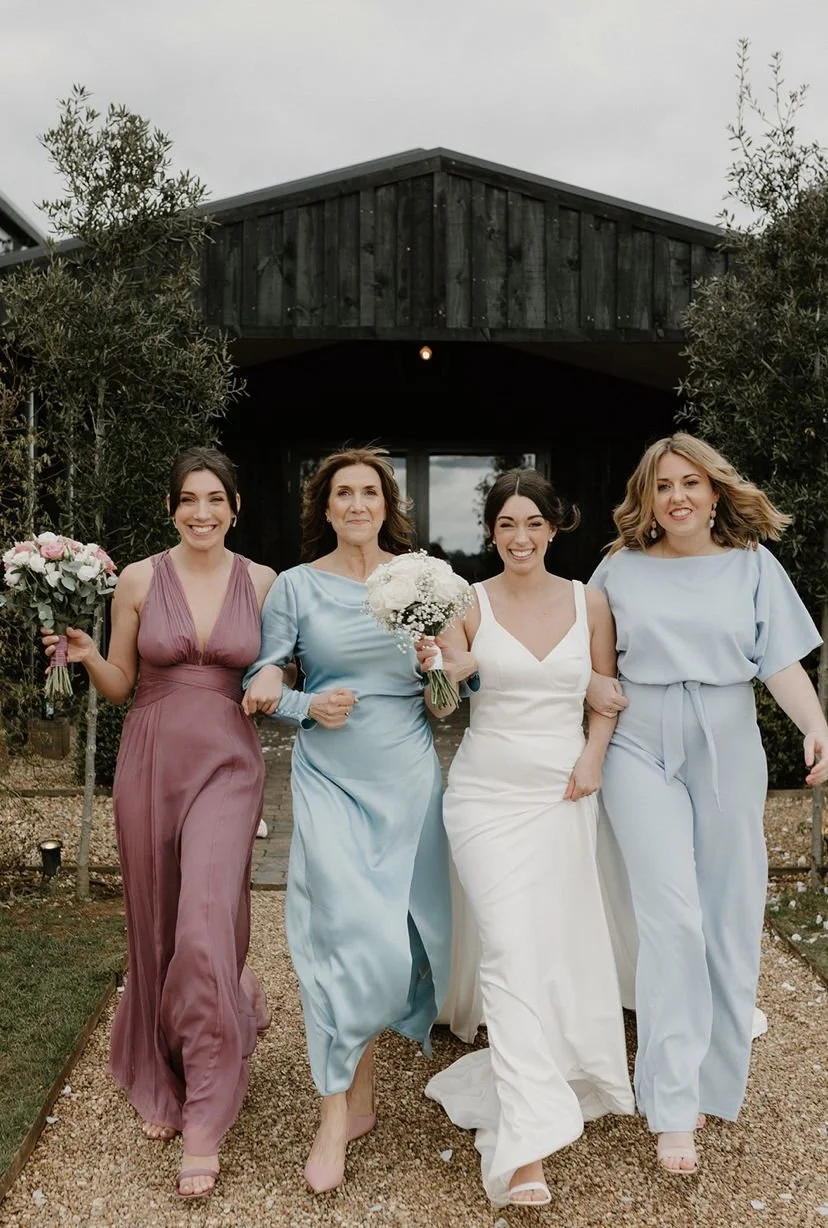 Four women, including a bride in a white wedding dress, walking outside on a gravel path, holding bouquets, with trees and a black wooden building in the background.