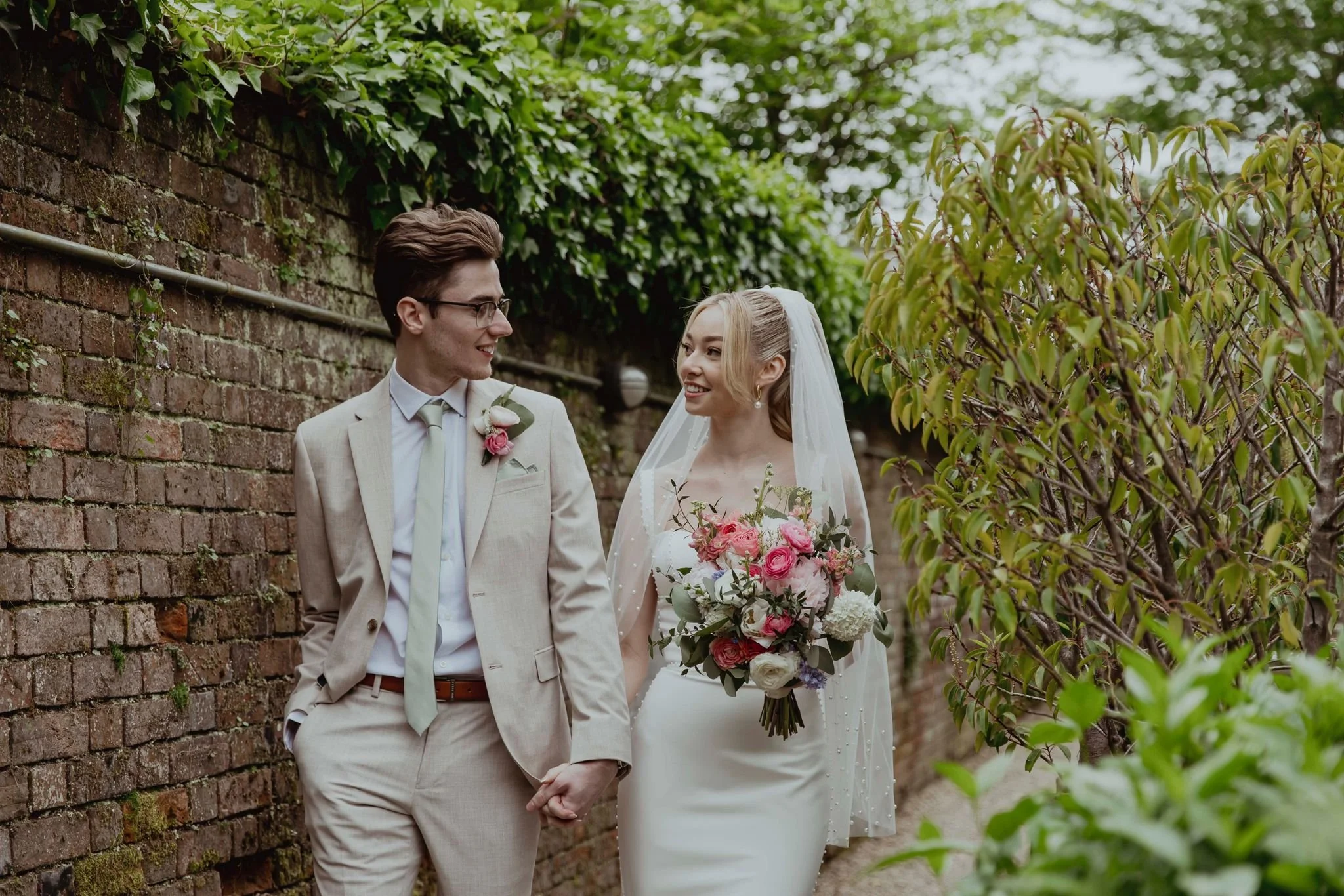 A wedding couple walking hand in hand outdoors next to a brick wall, with the bride holding a bouquet of pink and white flowers, both smiling at each other.