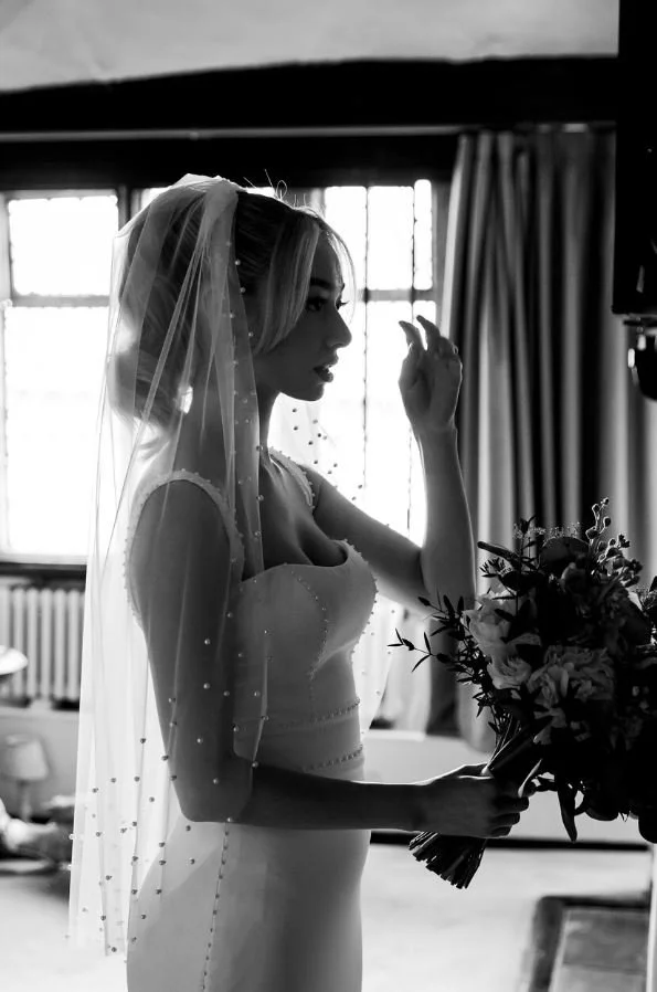 A black and white photo of a bride holding a bouquet and gazing out a window, wearing a wedding dress and veil.