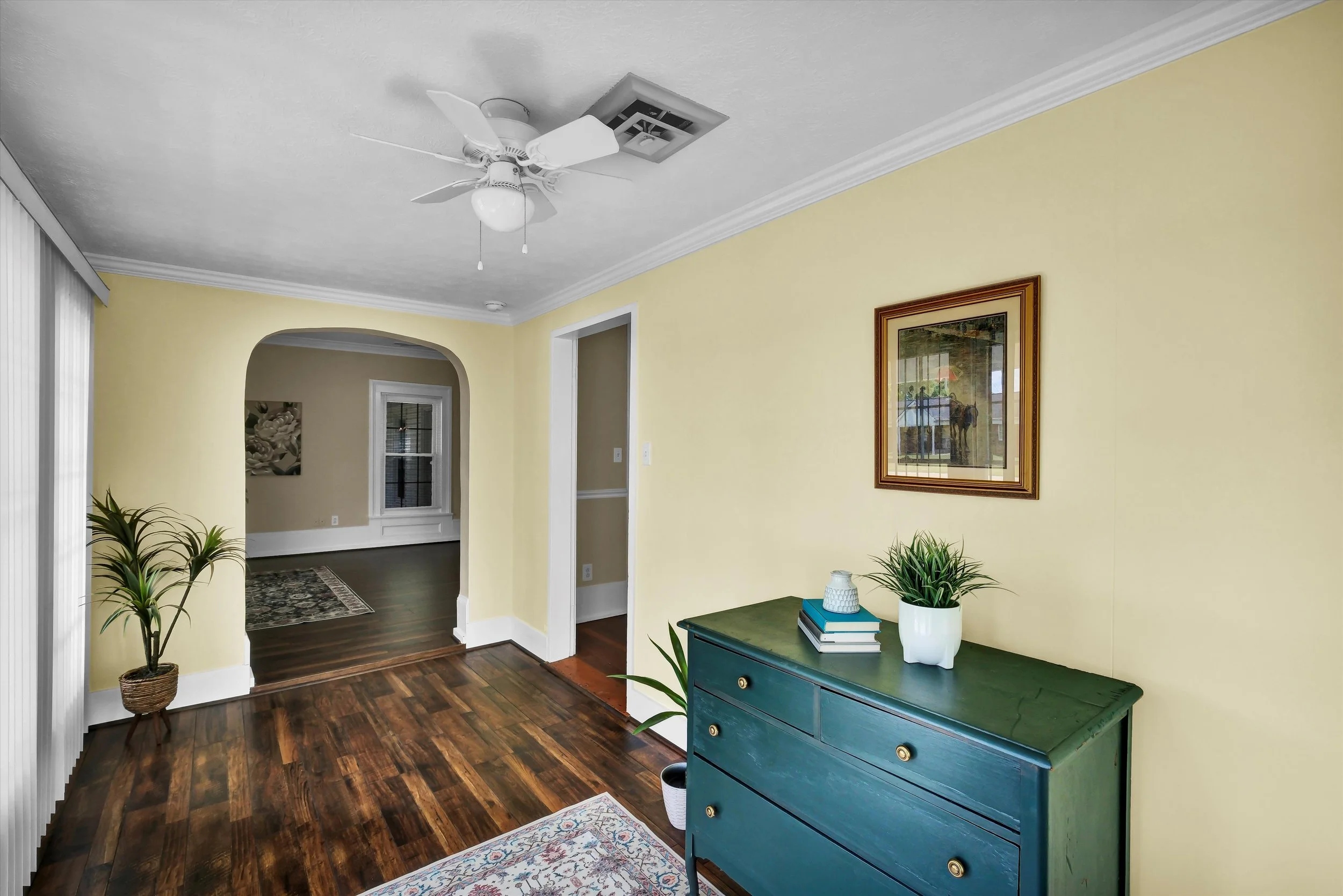 Interior of a home with yellow walls, dark wood flooring, a green dresser with decorative items, a framed picture on the wall, a ceiling fan, a window with white trim, and an adjoining room with a rug and window.