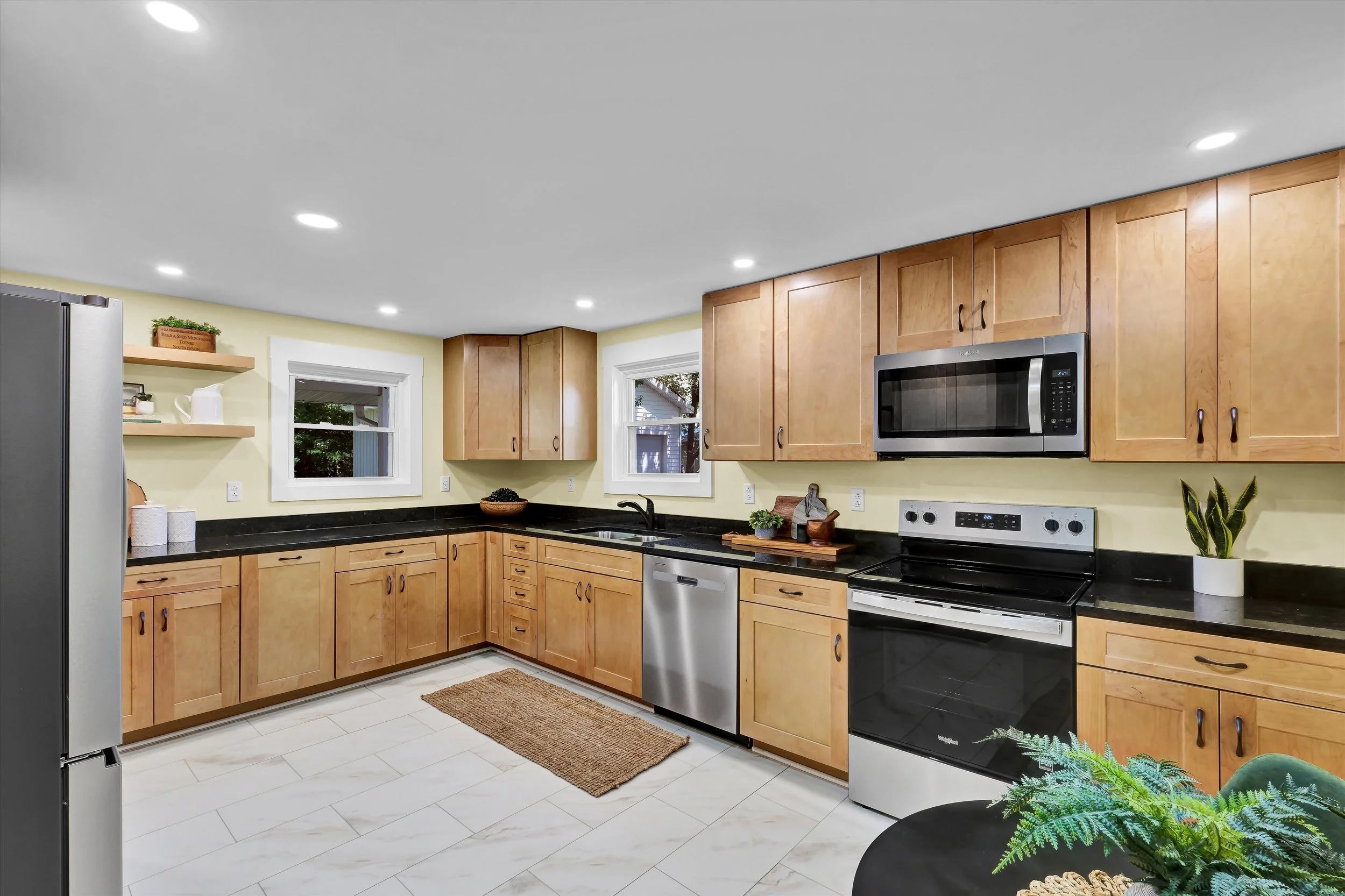 Modern kitchen with wooden cabinets, black countertops, stainless steel appliances, and a tiled floor. There are two small windows above the sink, and decorative plants on the counter.
