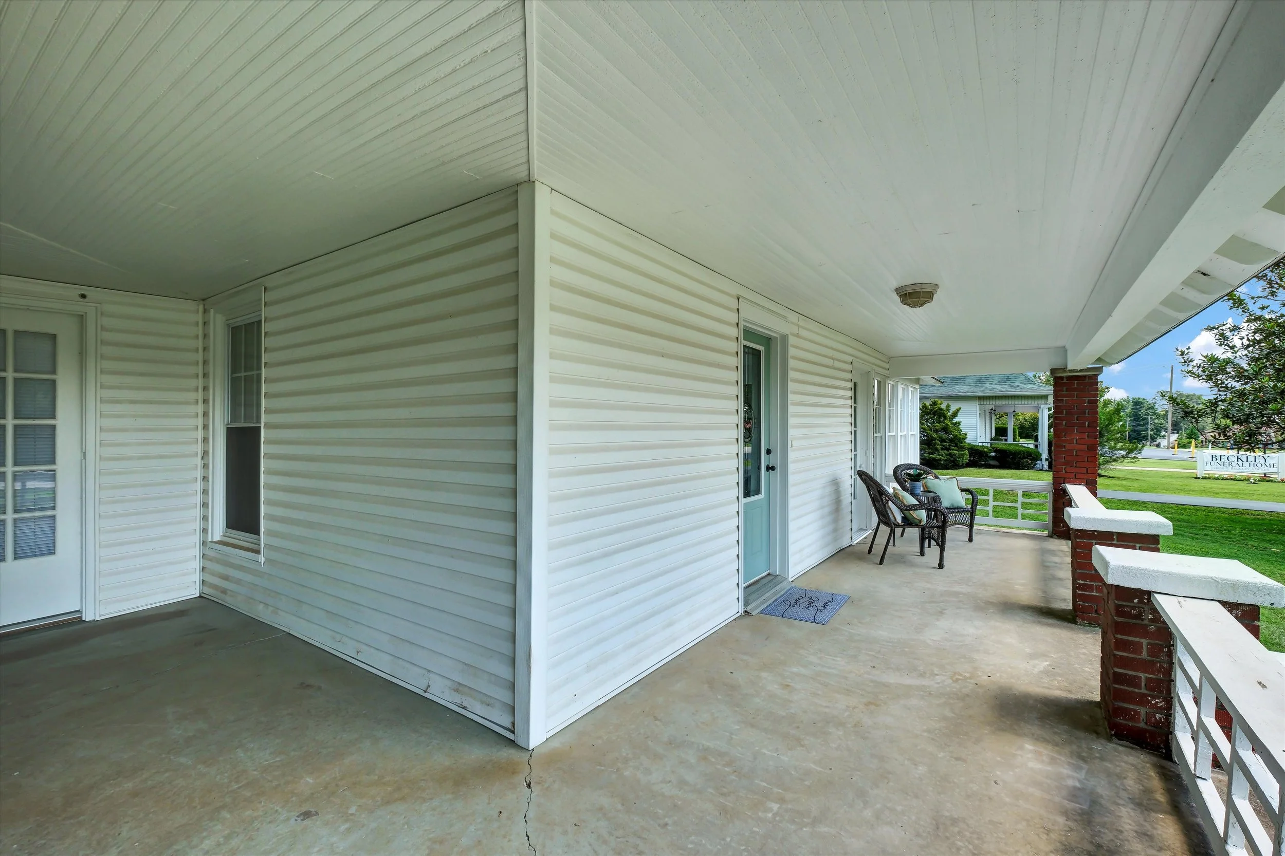 Image of a porch with white siding walls, a ceiling, and concrete floor. There are two wicker chairs with cushions, a small table, and a door leading inside. The porch is supported by brick pillars and overlooks a green yard with trees and a sign in 