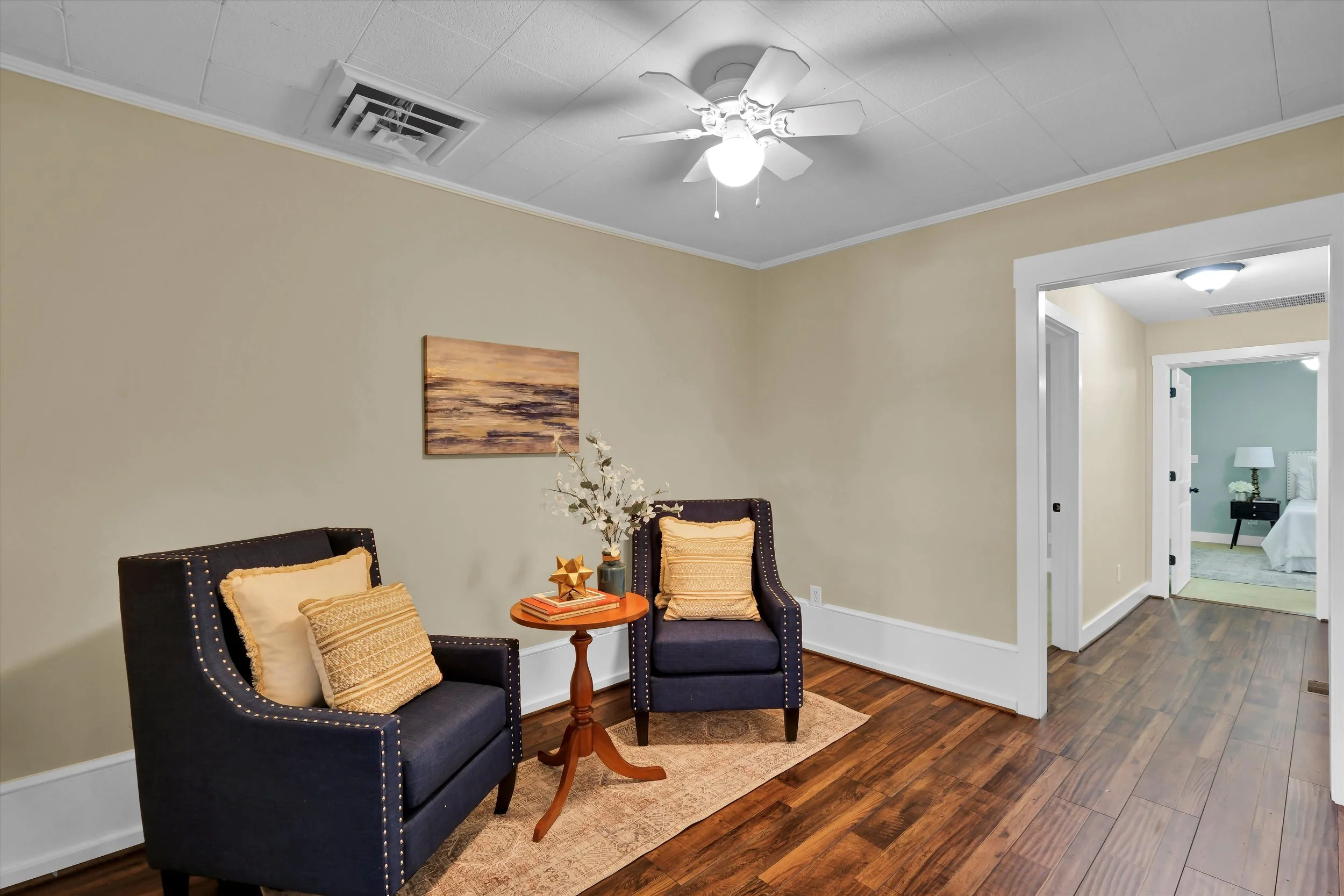 A cozy living room with two dark blue armchairs with beige pillows, a small wooden side table with decor, a beige wall with a landscape painting, wooden flooring, and a ceiling fan with a light fixture. There is an open doorway leading to a bedroom w