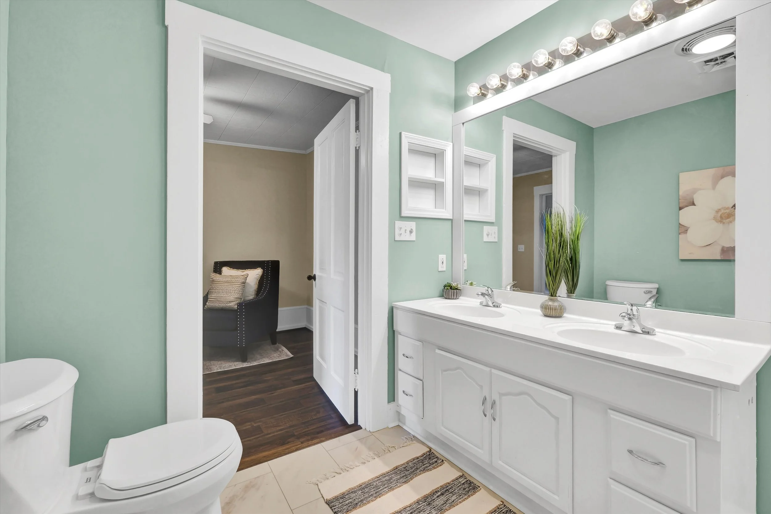 Bathroom with double sink vanity, large mirror, light green walls, potted plants, and a door leading to a room with a black armchair, beige walls, and dark hardwood floors.