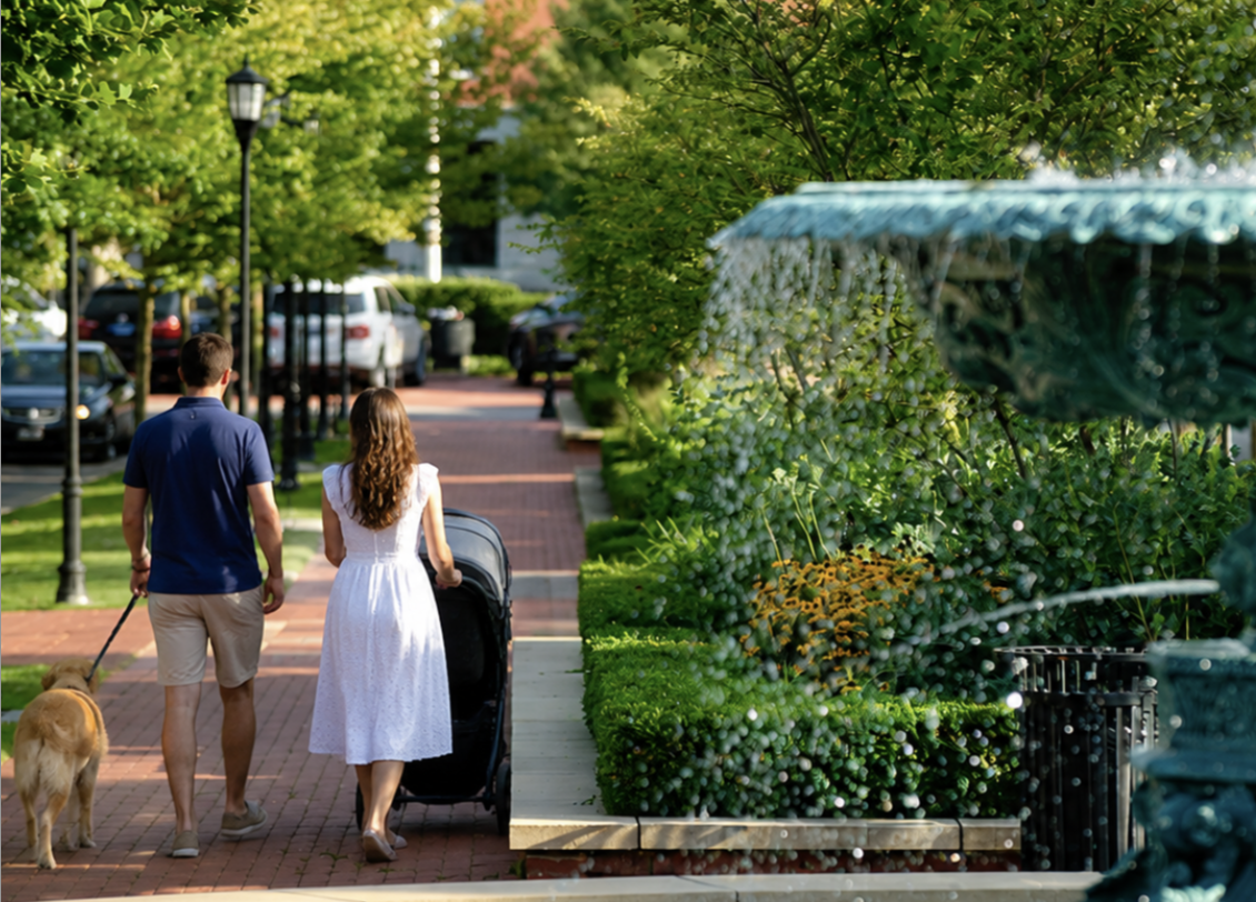 A couple walking down a brick sidewalk in Louisville with a large dog, surrounded by green trees and bushes in a neighborhood with parked cars.