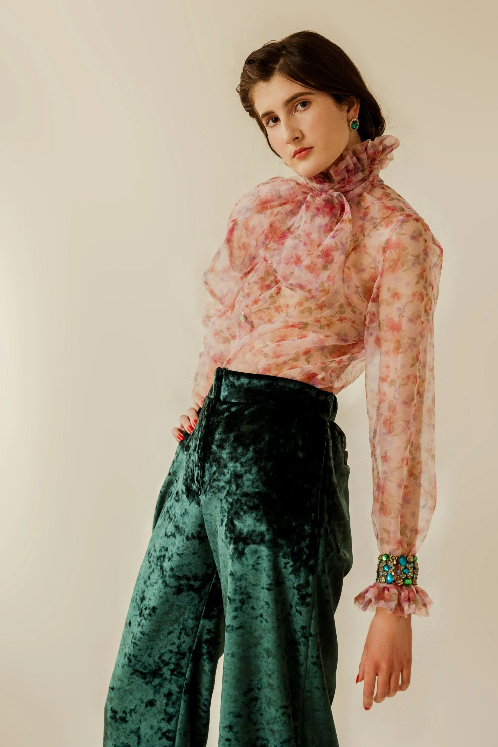 A woman wearing a sheer floral blouse with ruffled collar and cuffs, and high-waisted velvet pants, standing against a plain background.