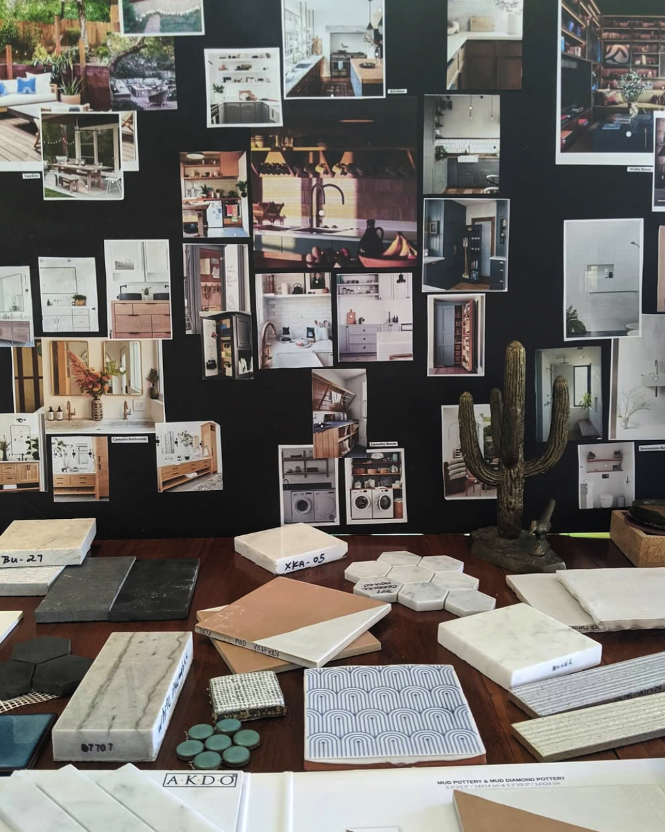 A display board with various interior design photos of kitchens, bathrooms, and living spaces, alongside samples of tiles, stones, and materials on a table in front, including cacti and a small fabric sample.
