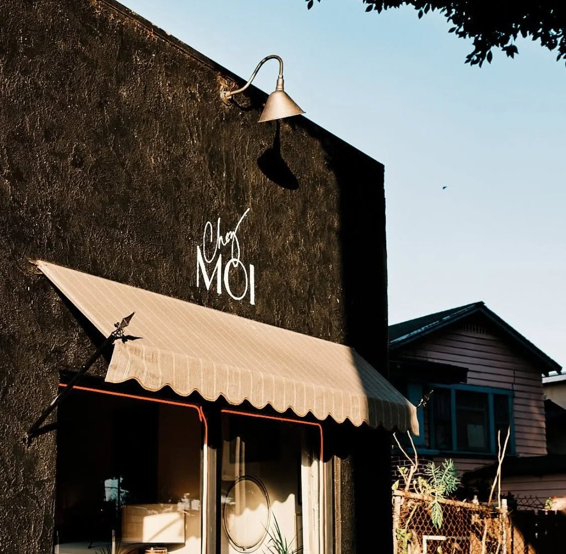 Exterior of Shop Chez Moi building with a beige awning above a glass window, black textured wall, and a silver outdoor light fixture.  Curated Vintage Furniture, Bespoke Furniture and Gallery
