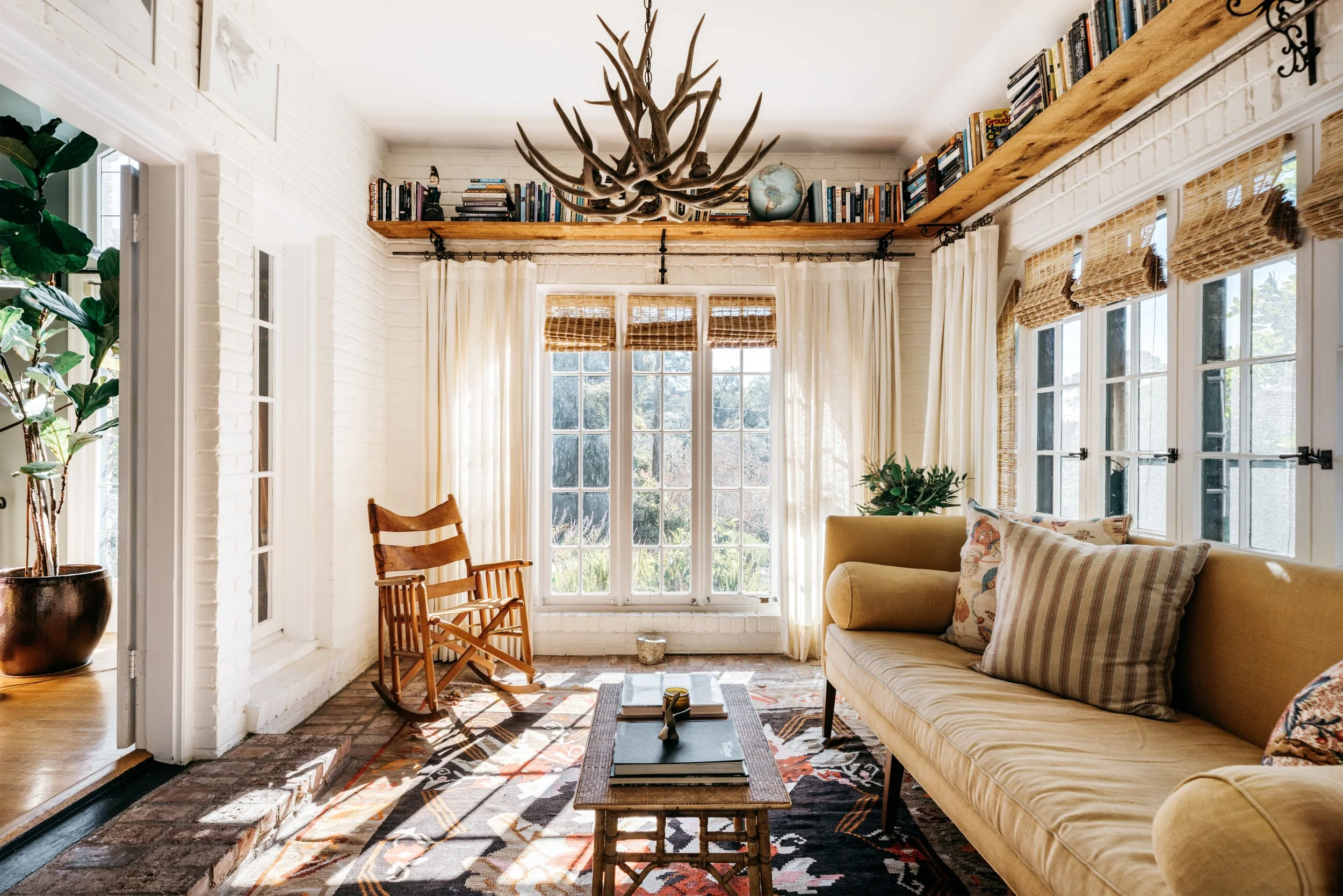 Bright living room with large windows, white brick walls, cream curtains, wooden bookshelf above window, vintage rocking chair, beige sofa with pillows, potted plant, patterned rug, and a rustic ceiling light fixture.