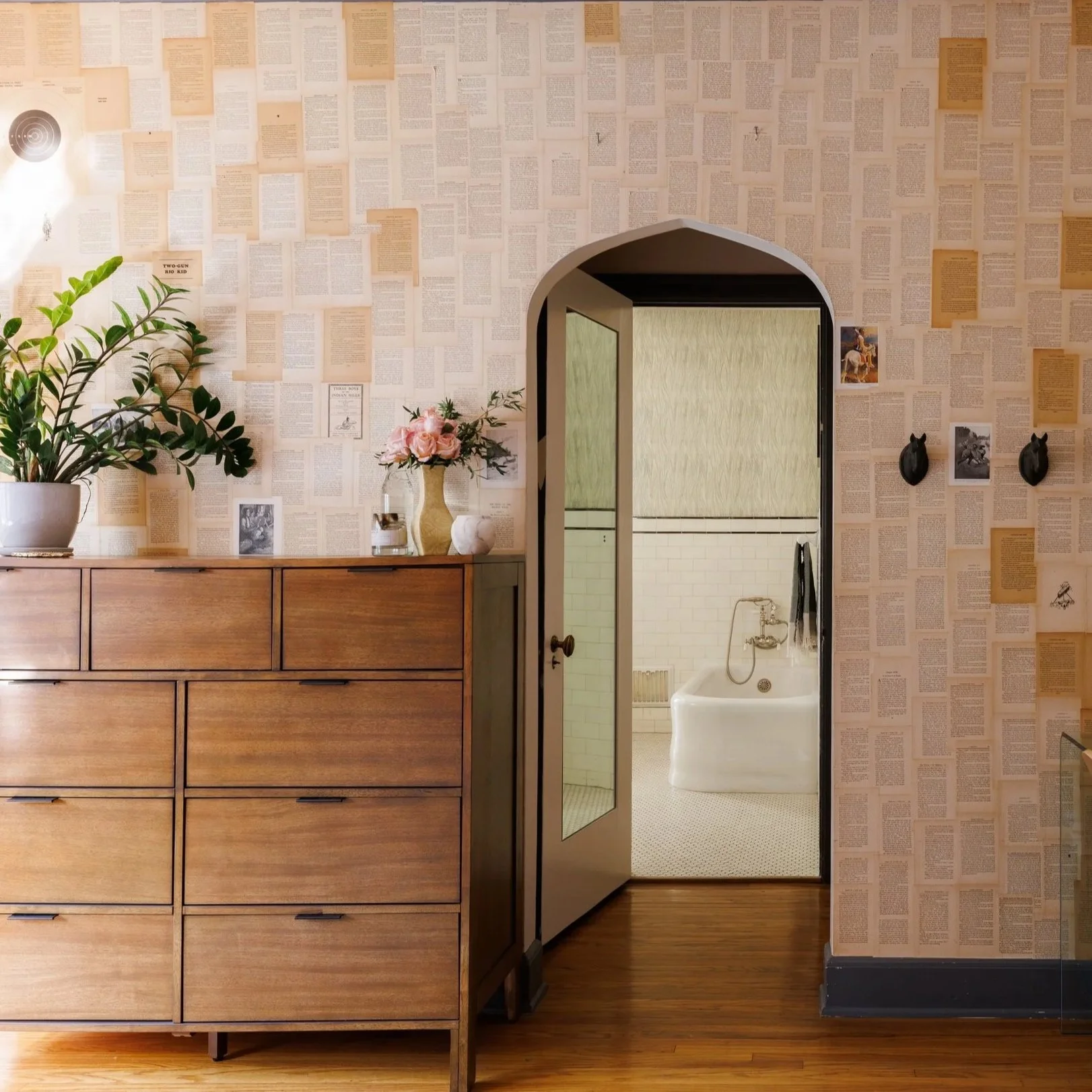 Interior decor of a Childs room with wallpaper made of printed pages, a wooden dresser with potted plants and flowers on top, and a door leading to a bathroom with a bathtub.