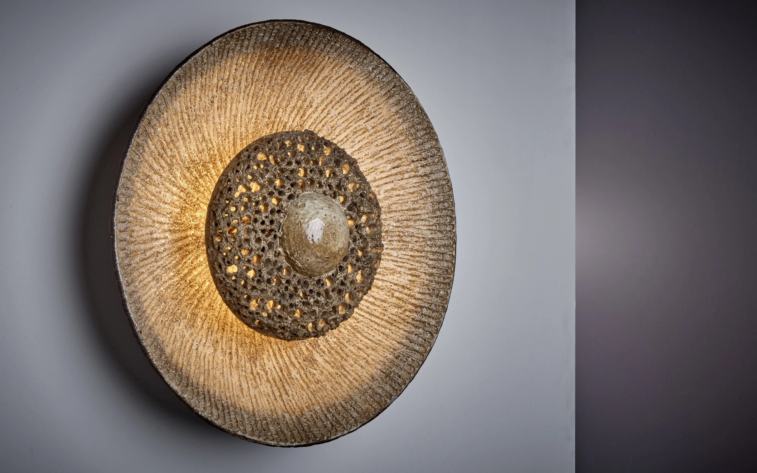 A decorative ceramic wall lamp with a circular design featuring a large outer ring with textured lines, an inner ring with a carved pattern, and a spherical element at the center, mounted on a partial wall with soft ambient light.