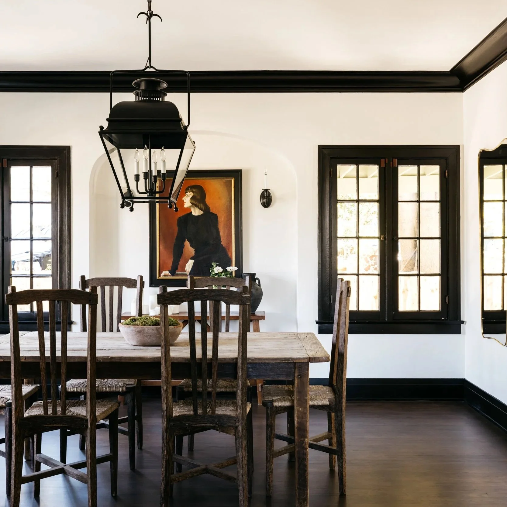 A dining room with a wooden table and six wooden chairs, black-trimmed windows, a hanging black lantern with candle-like lights, a painting of a woman in black on the wall, and a small mirror on the wall.