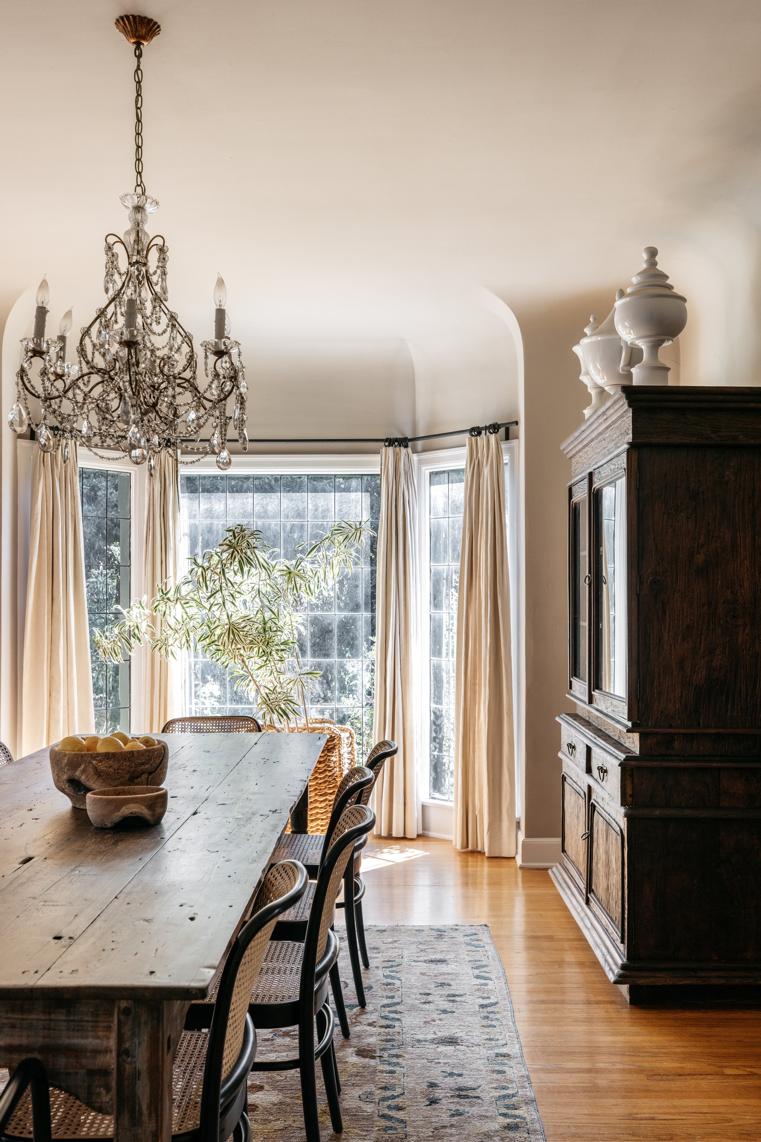 A dining room with a large wooden table, black chairs, a chandelier, a tall dark wood cabinet, large windows with cream curtains, a potted plant, and a patterned rug on a wooden floor. Mariah Obrien Interior Design Project
