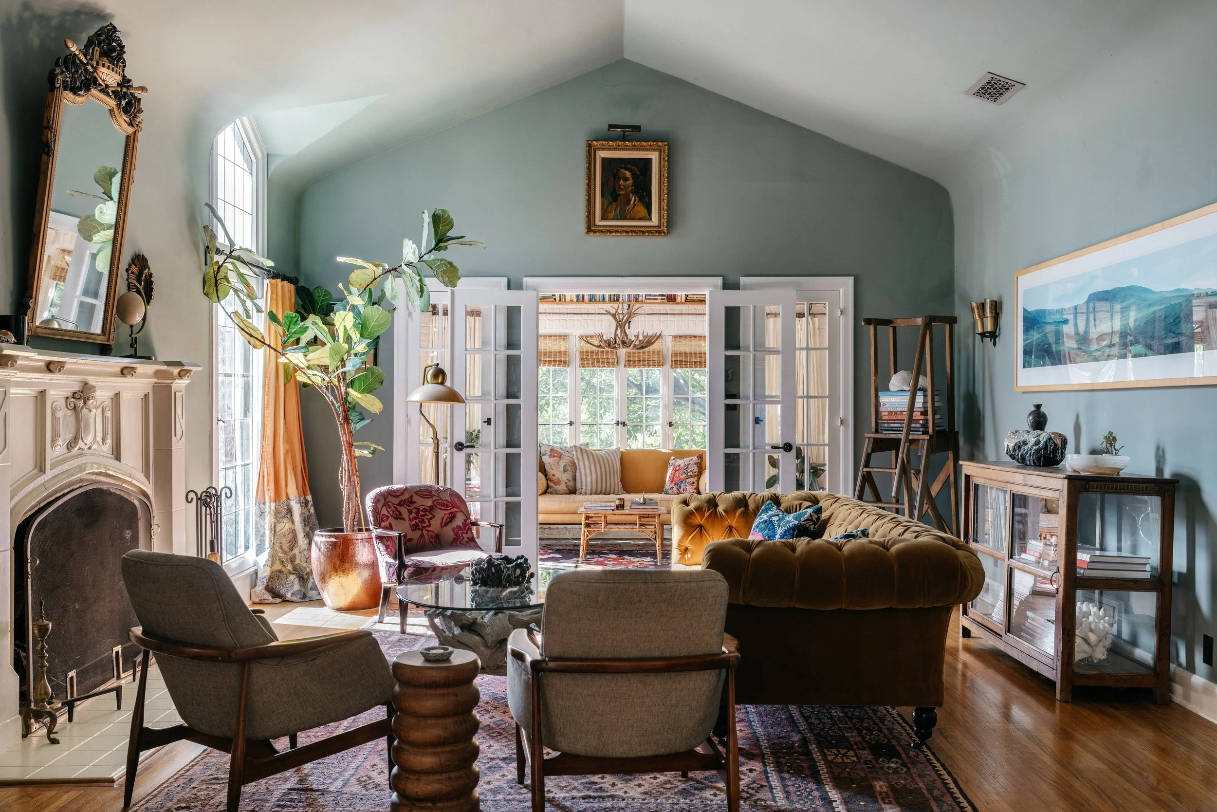 A vintage-style living room with a fireplace, a large window, and French doors leading to a sunroom. The room has eclectic furniture, including an olive green velvet sofa, armchairs, a glass coffee table, and a large potted plant. There are decorative paintings, a large mirror, and various artworks and books, creating a cozy and stylish ambiance.