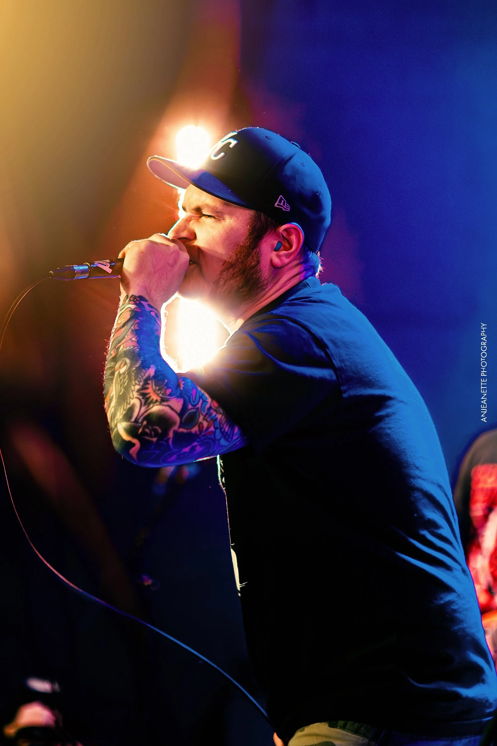 Male singer with tattoos on his arm singing into a microphone, wearing a baseball cap and a black shirt, with colorful stage lighting.