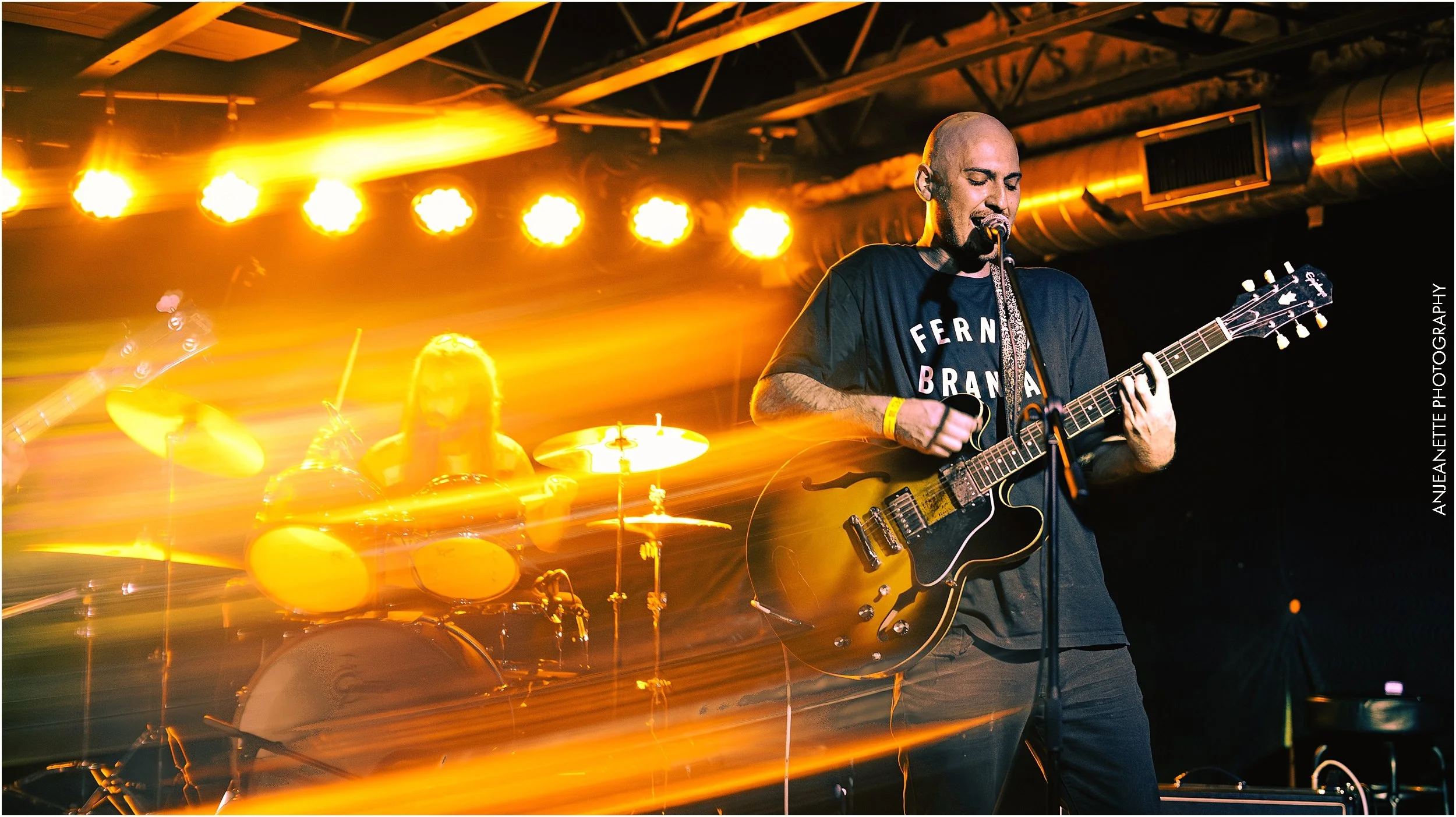 Musician playing an electric guitar singing into a microphone during a live concert, with a drummer in the background and orange stage lights creating motion blur.