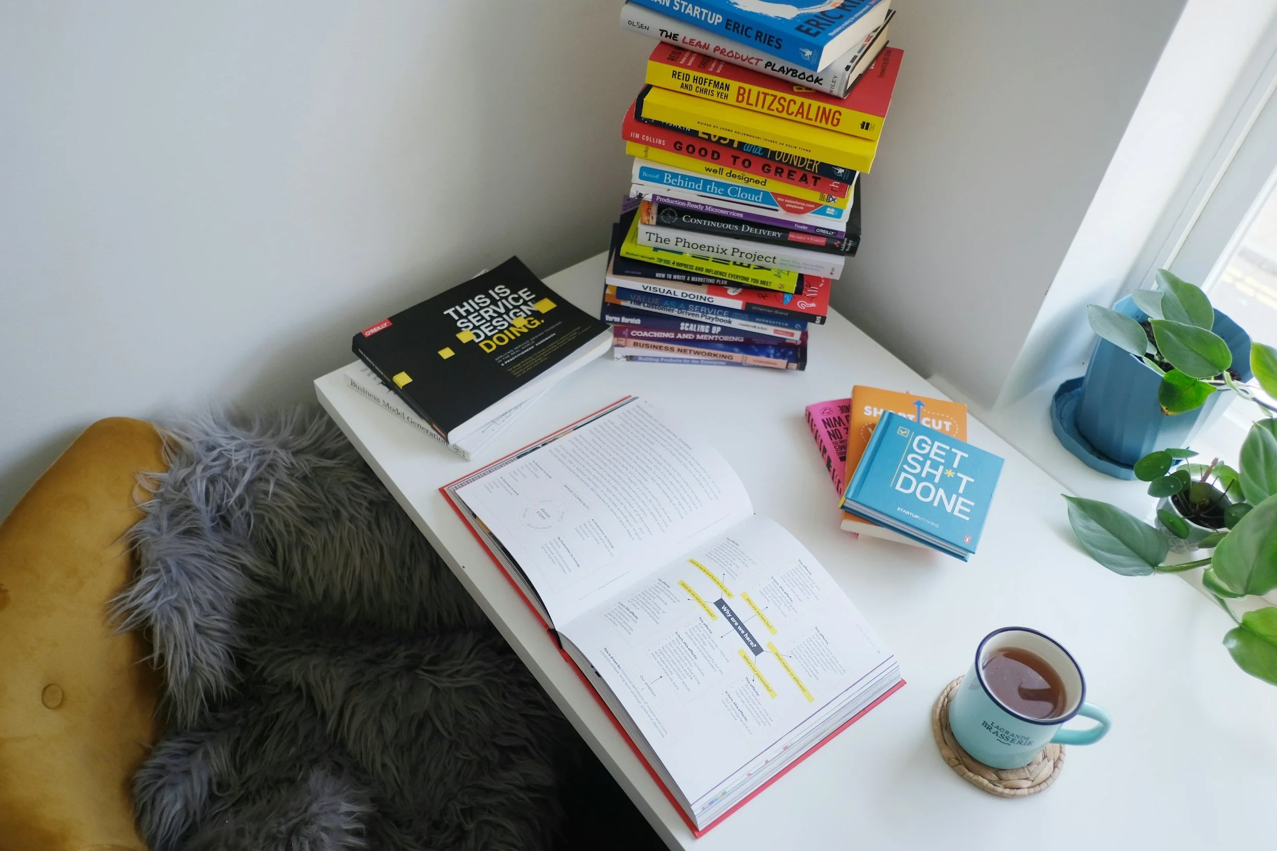 A white desk with a stack of colorful books, open notebook, a turquoise coffee mug on a coaster, and a blue potted plant next to a window. Furry blanket draped over a yellow chair is visible in the lower left corner.