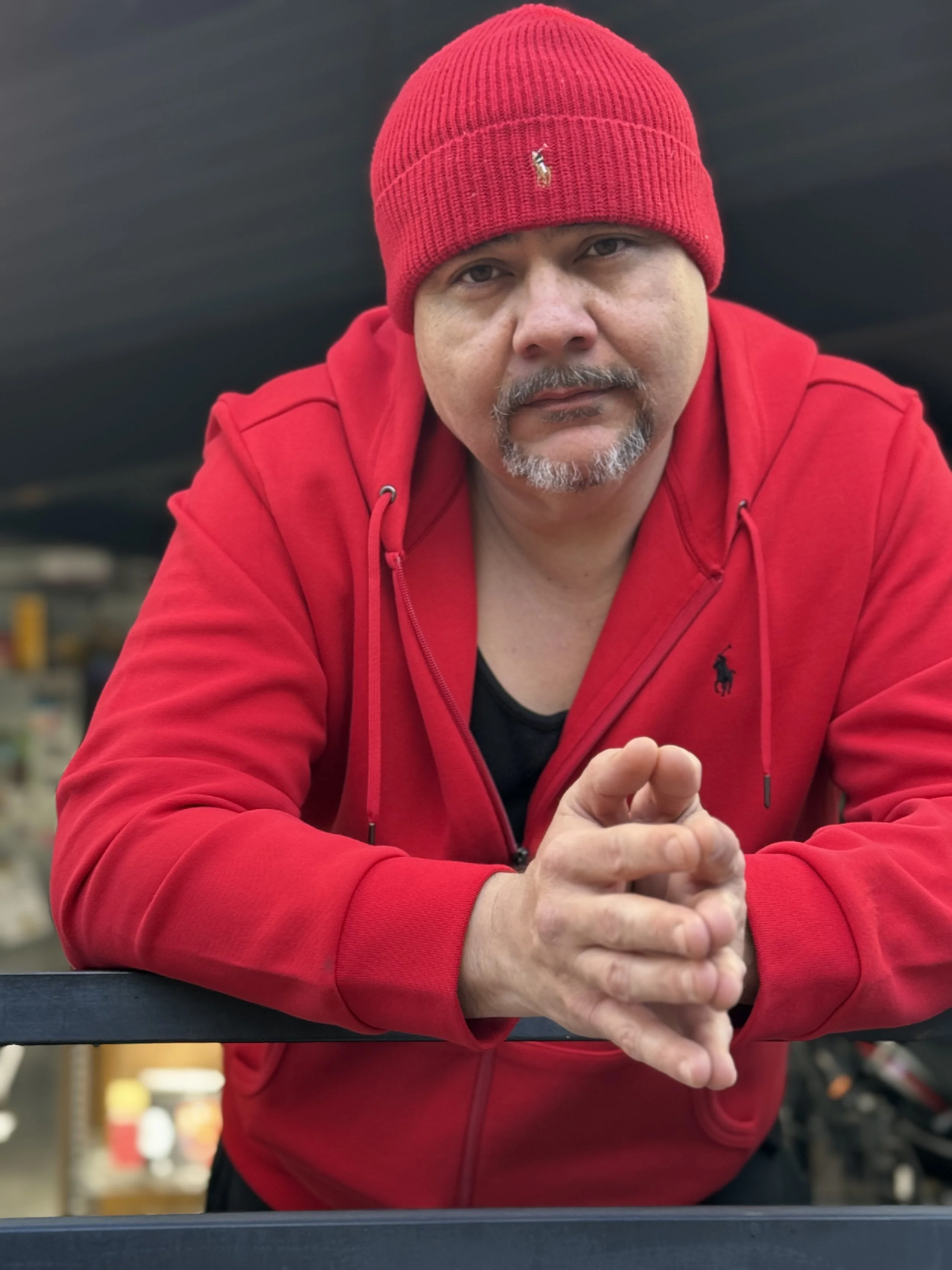 A man with a beard and mustache wearing a red beanie and red jacket, leaning forward with his hands clasped, looking at the camera.