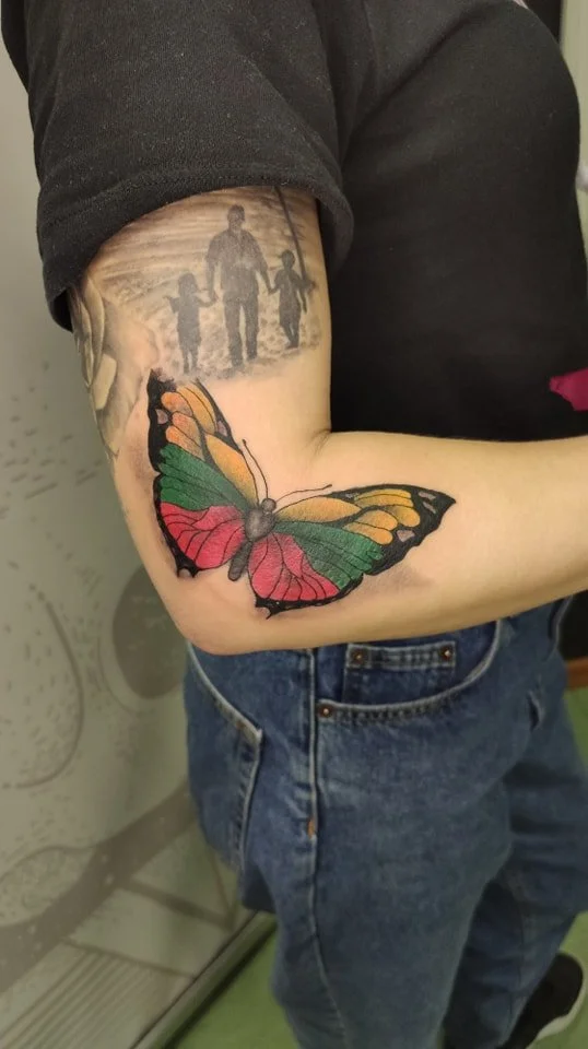 A person with a black t-shirt and blue jeans showing a colorful butterfly tattoo on their arm and a grayscale family portrait tattoo on the upper arm.