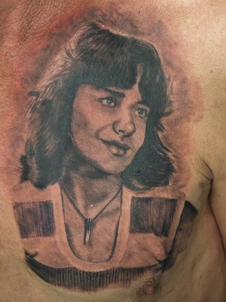 A detailed black and gray tattoo of a young woman with shoulder-length hair and a necklace, tattooed on a person's chest.