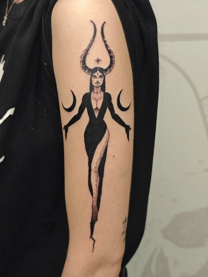 Tattoo of a stylized woman with horns, a crescent moon on either side of her, and a star above her forehead on upper arm.