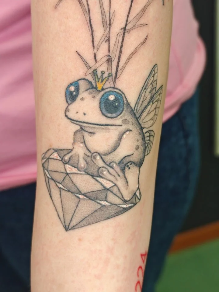 A tattoo of a cute frog with large eyes, a small crown, fairy wings, and thin antennae, sitting inside a diamond-shaped gem.