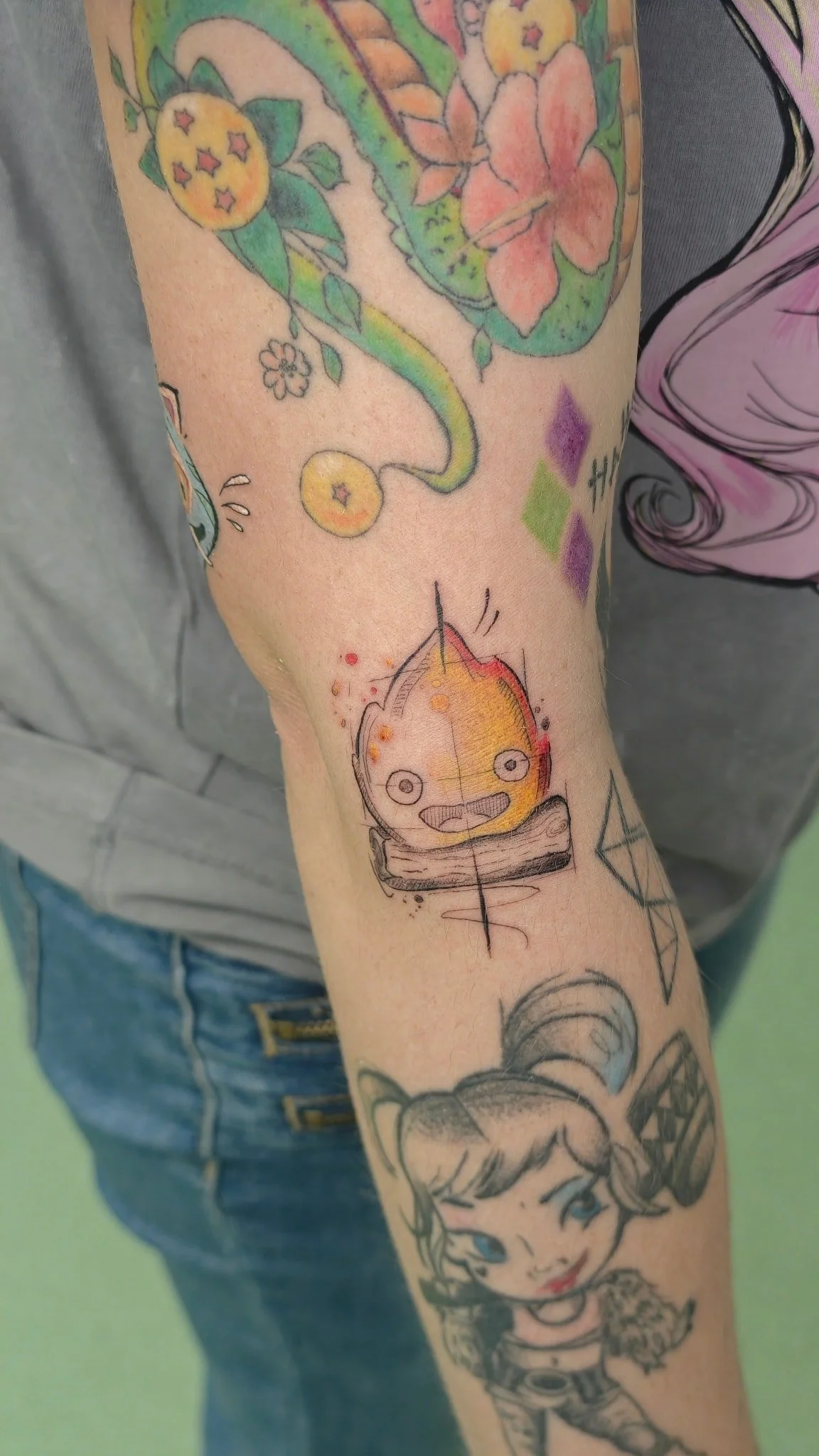 A collection of colorful tattoos on a person's arm, including a smiling cartoonish creature with large eyes, a branch with a flower, and various abstract designs.
