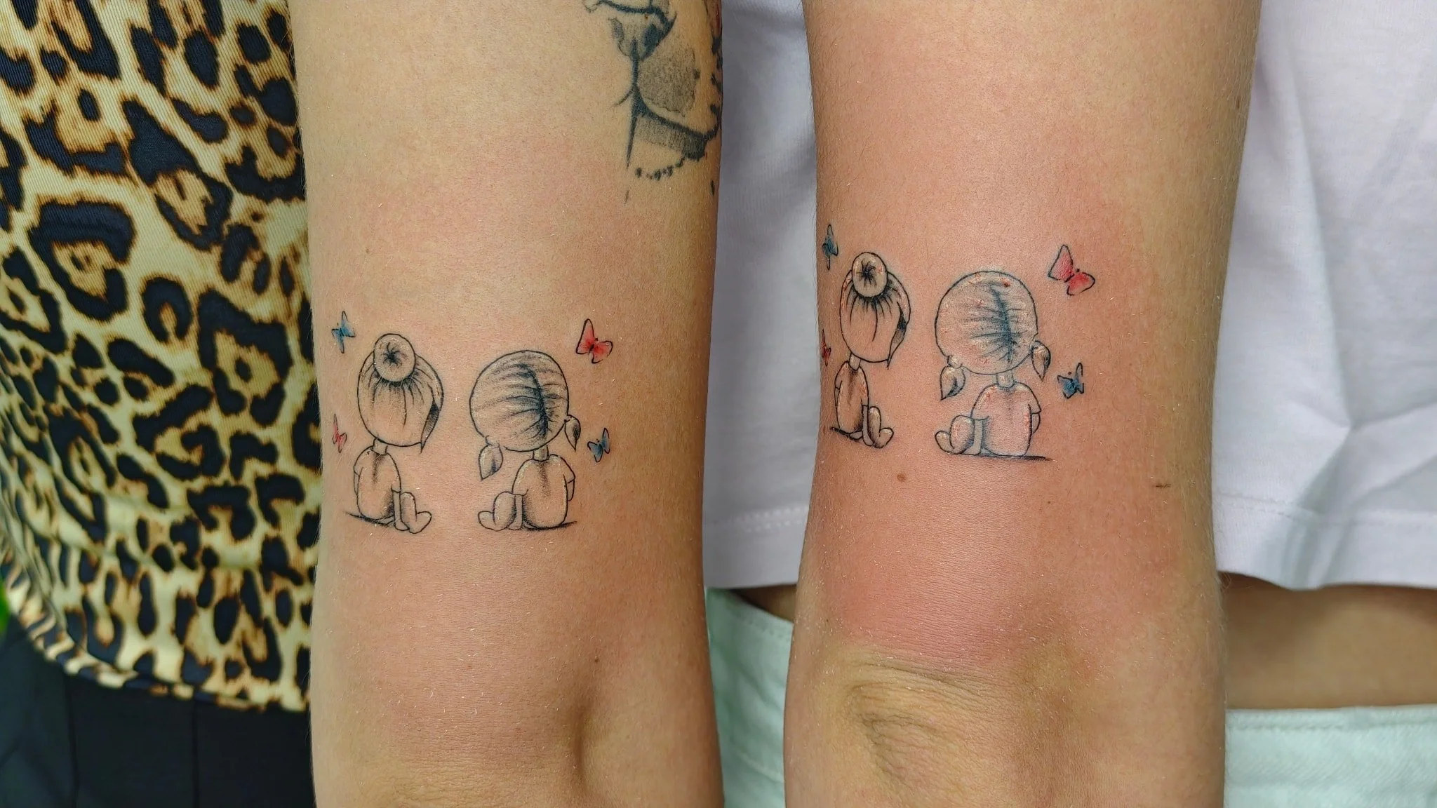 Two people have matching tattoos of a girl with pigtails sitting on the ground, surrounded by small colorful butterflies.