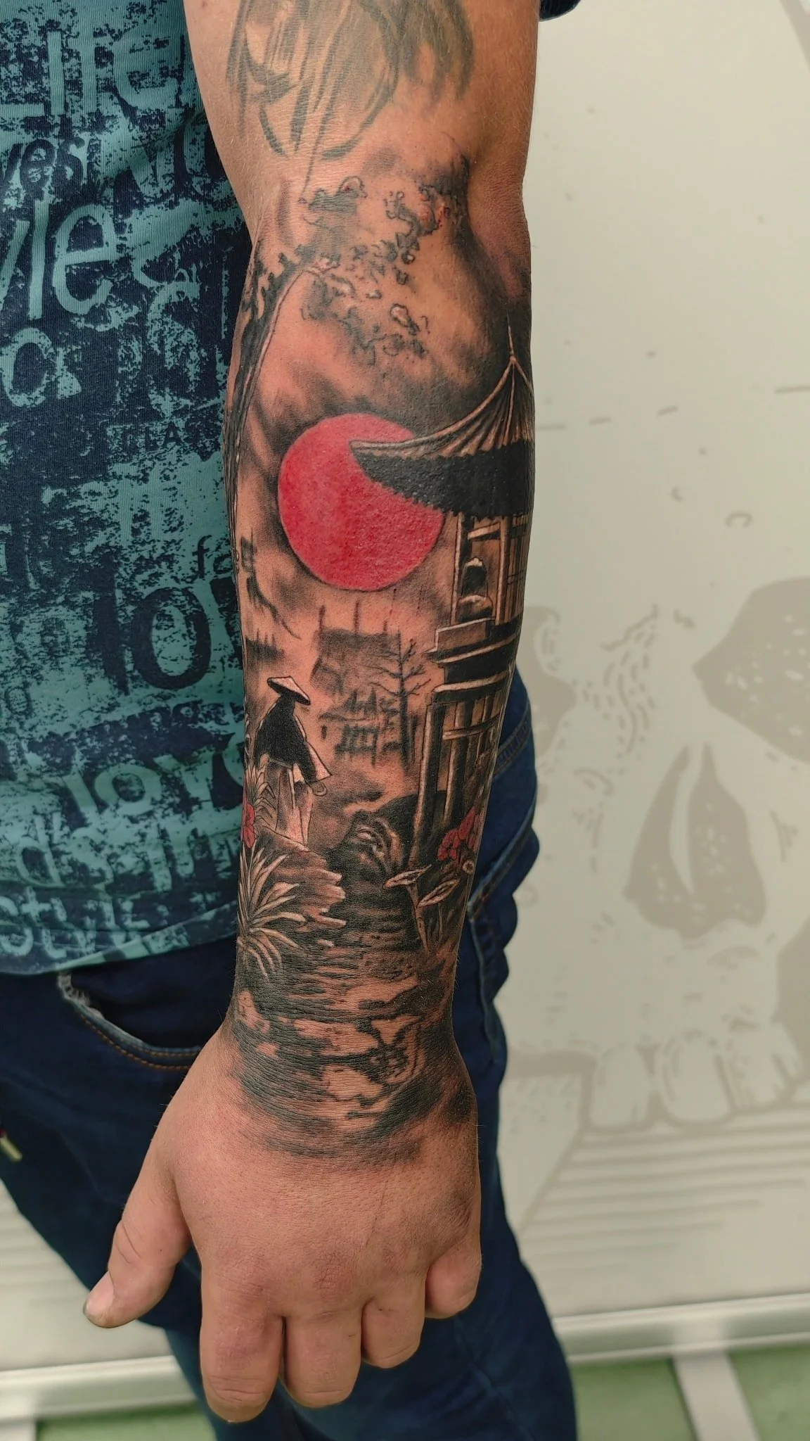 Tattoo of a Japanese landscape featuring a red sun, a pagoda, a bridge, a person in traditional clothing, and trees.