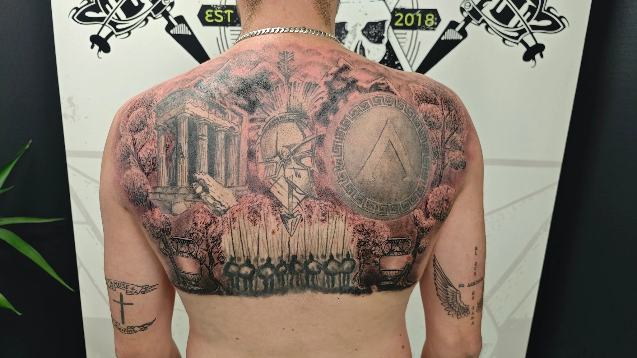 Tattooed back of a person featuring ancient ruins, a compass, Greek key border, fire, trees, an ancient coin with an arrow symbol, and a group of people holding flags, with additional tattoos on the arms of a cross and a winged heart with the words "