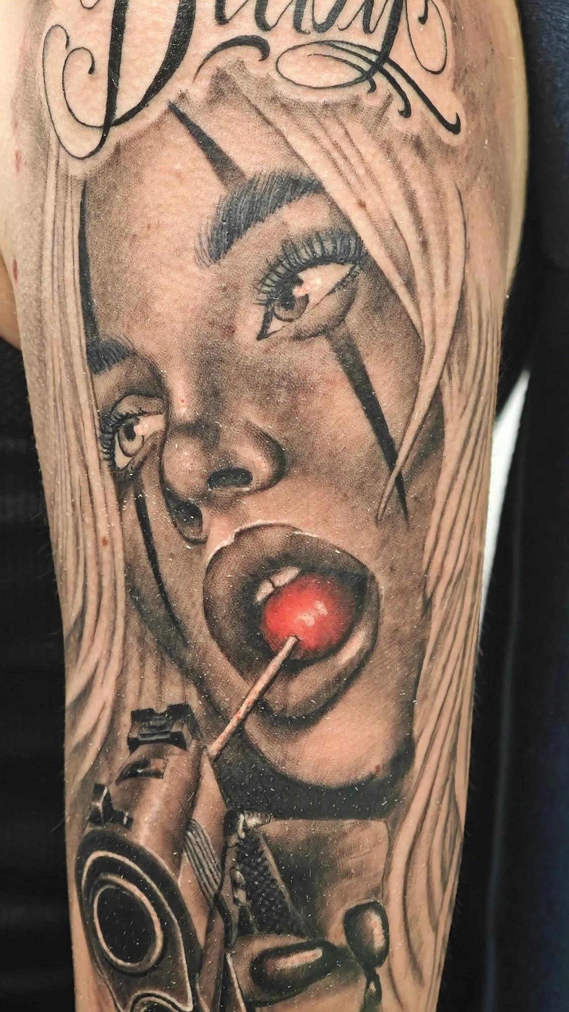 A black and gray tattoo of a woman with blonde hair, bold makeup, and black tears down her face. She is holding a gun to her mouth with a red lollipop in her mouth. The tattoo includes detailed shading and realistic features.
