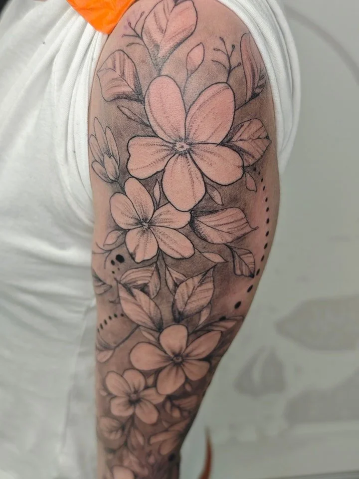 Black and gray floral tattoo featuring large and small flowers, leaves, and dotted designs on a person's upper arm and shoulder.