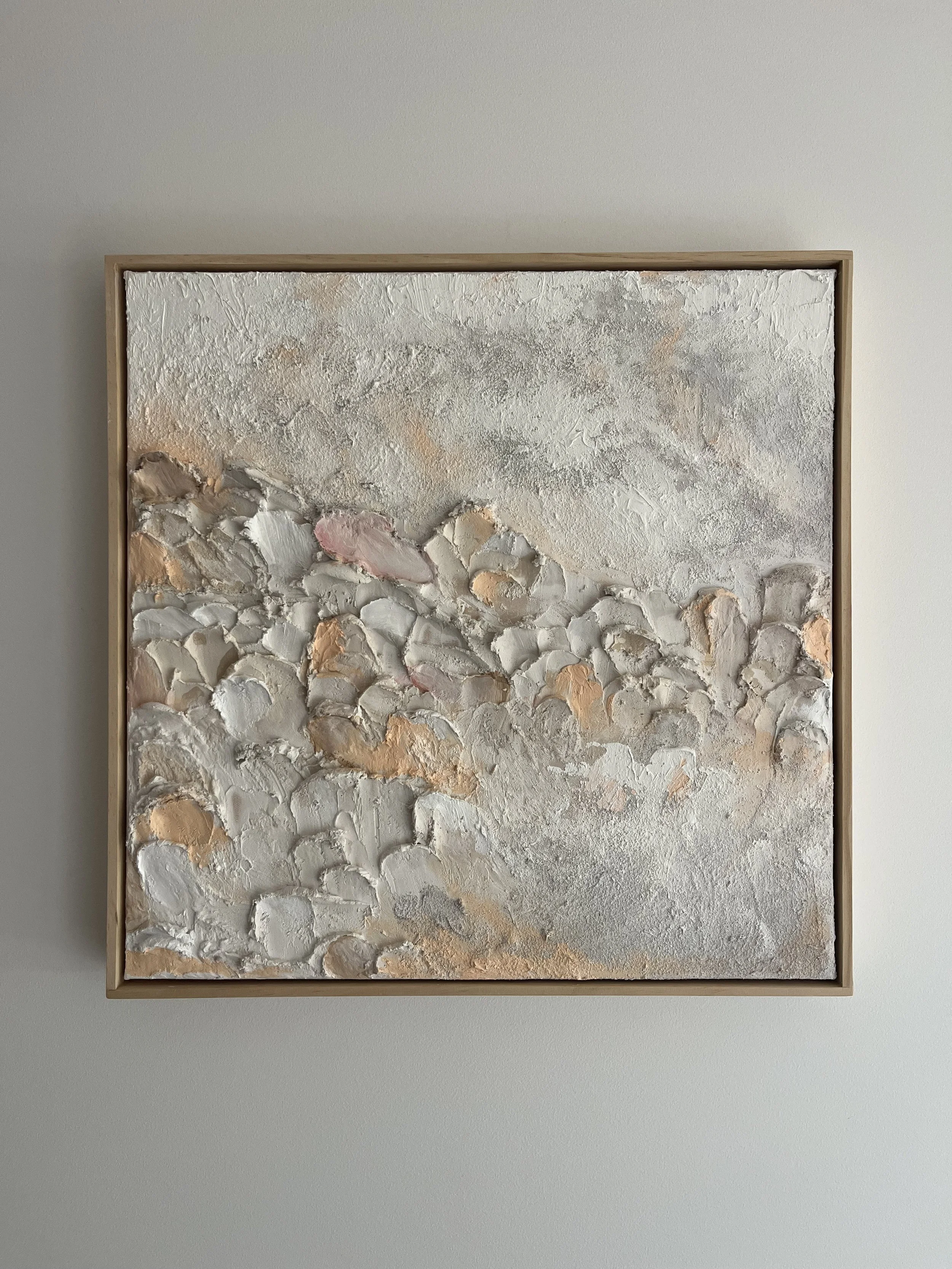 Abstract textured artwork with a mix of white, beige, pink, and gray hues, framed in light wood, hanging on a plain light-colored wall.