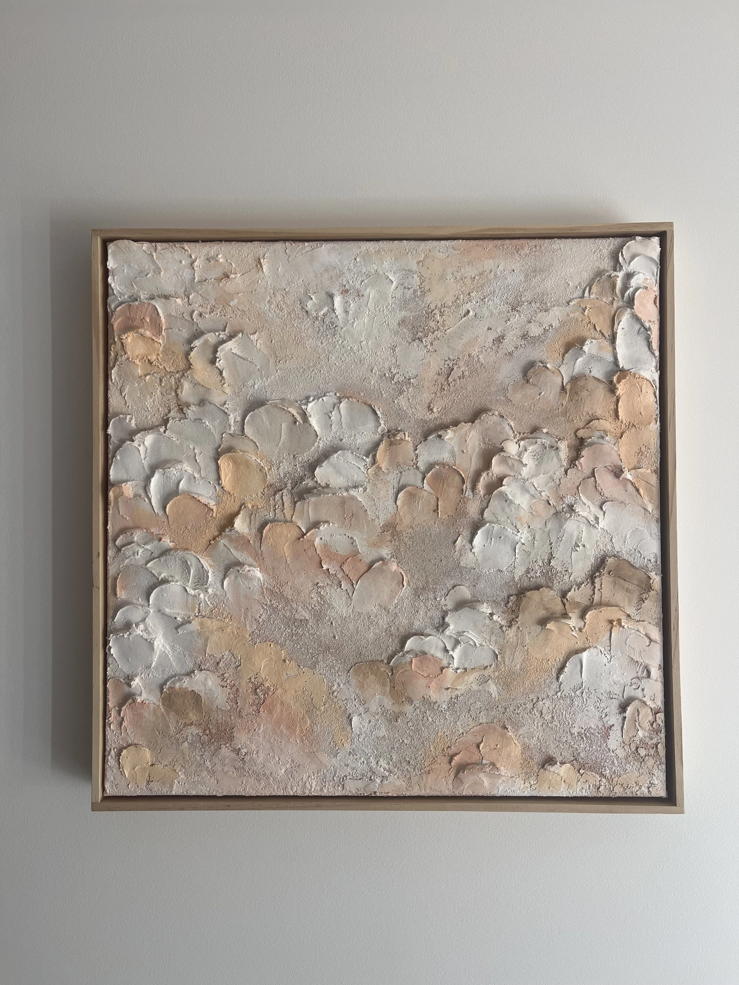 Abstract textured artwork with soft neutral colors framed in light wood, hung on a plain white wall.