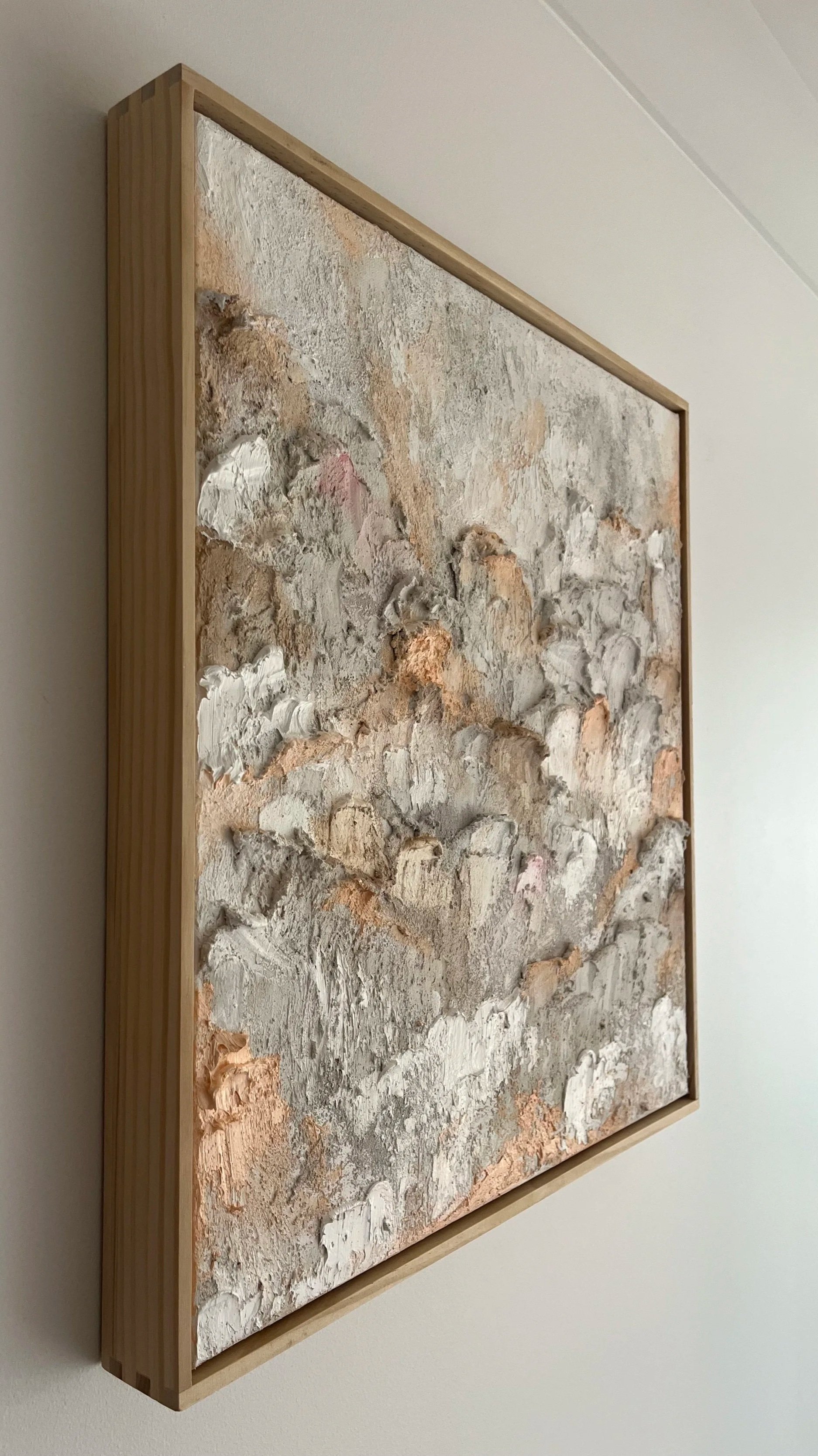 Mixed media artwork with textured, earthy-colored surfaces framed in light wood, displayed on a white wall.