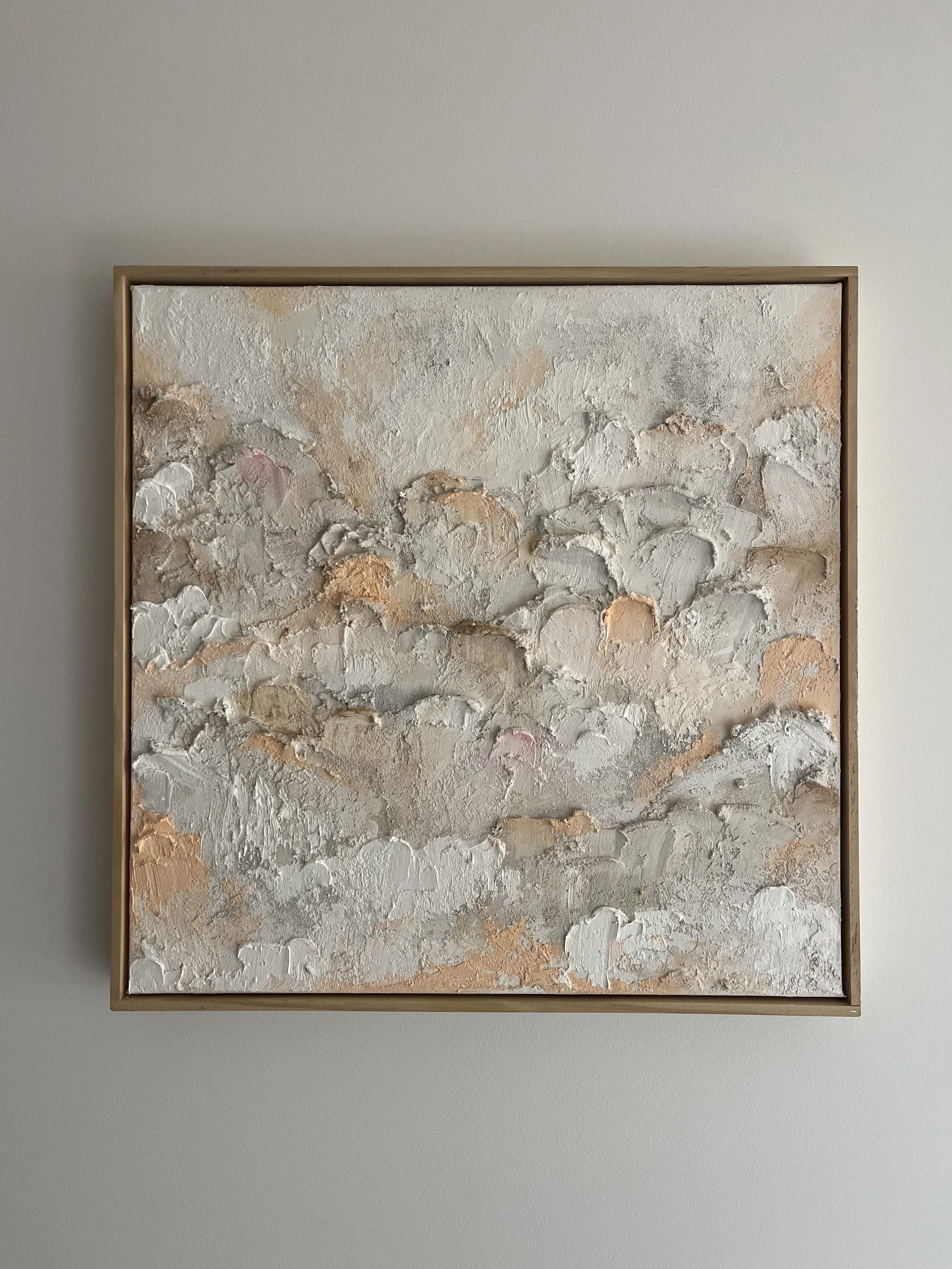 Abstract textured painting with white, beige, pink, and peach colors in a wooden frame hanging on a plain wall.