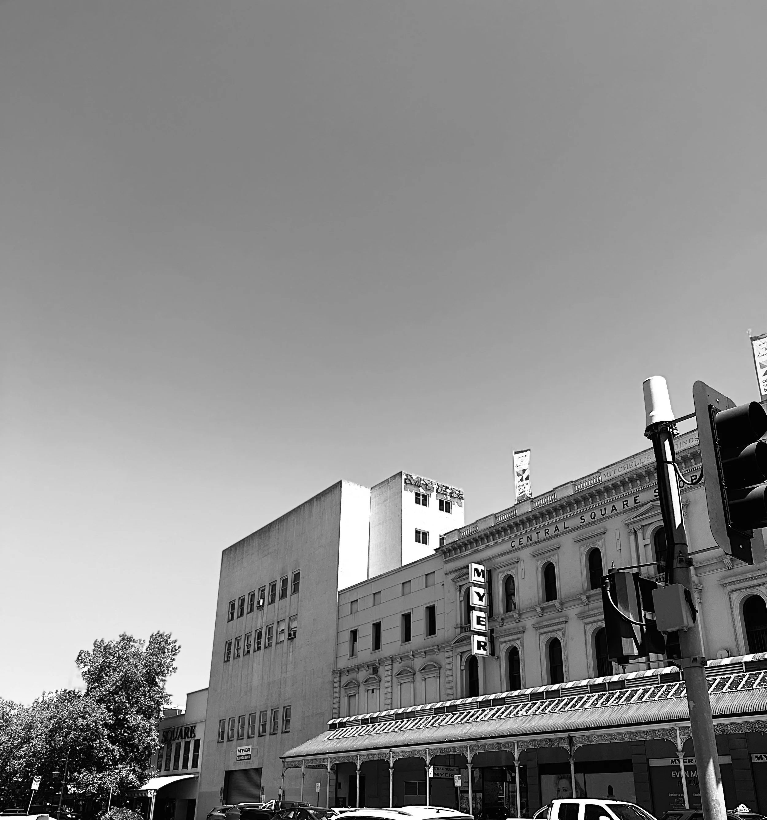 Black-and-white photo of buildings on a city street, with a large sign reading 'MYER' and a traffic light in the foreground.