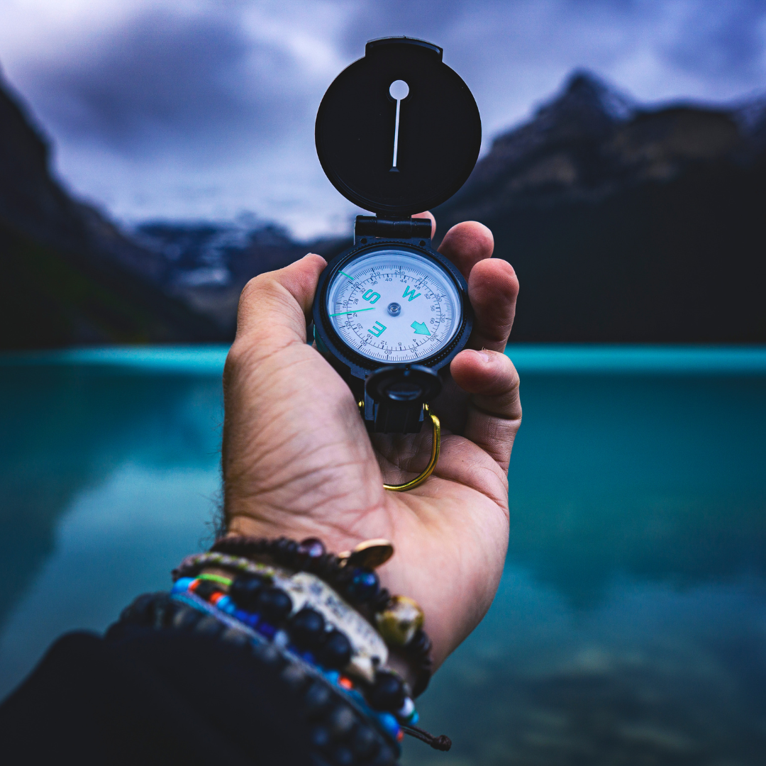 Hand holding a compass with a scenic mountain lake and cloudy sky in the background.