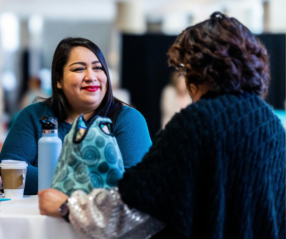 Two women sitting at a table talking; one woman is smiling, dressed in a blue sweater, with a water bottle, coffee cup, and a bag on the table.