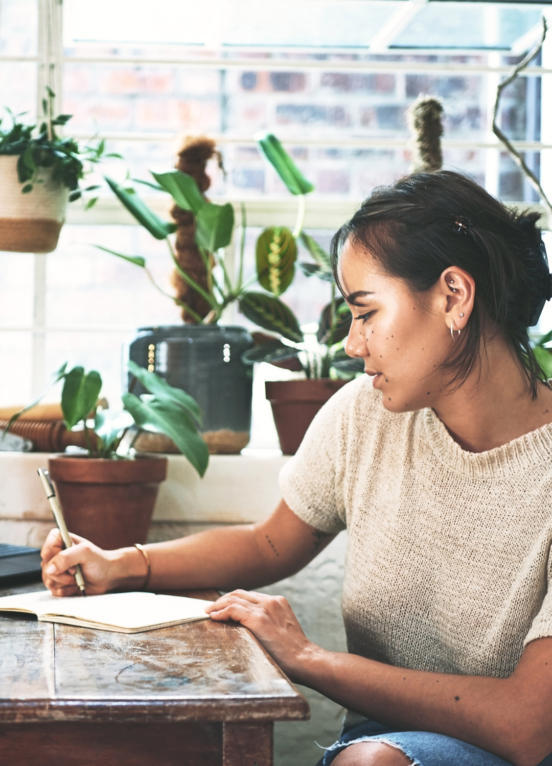 A woman sitting at a wooden table writing in a notebook, surrounded by potted plants by a window.