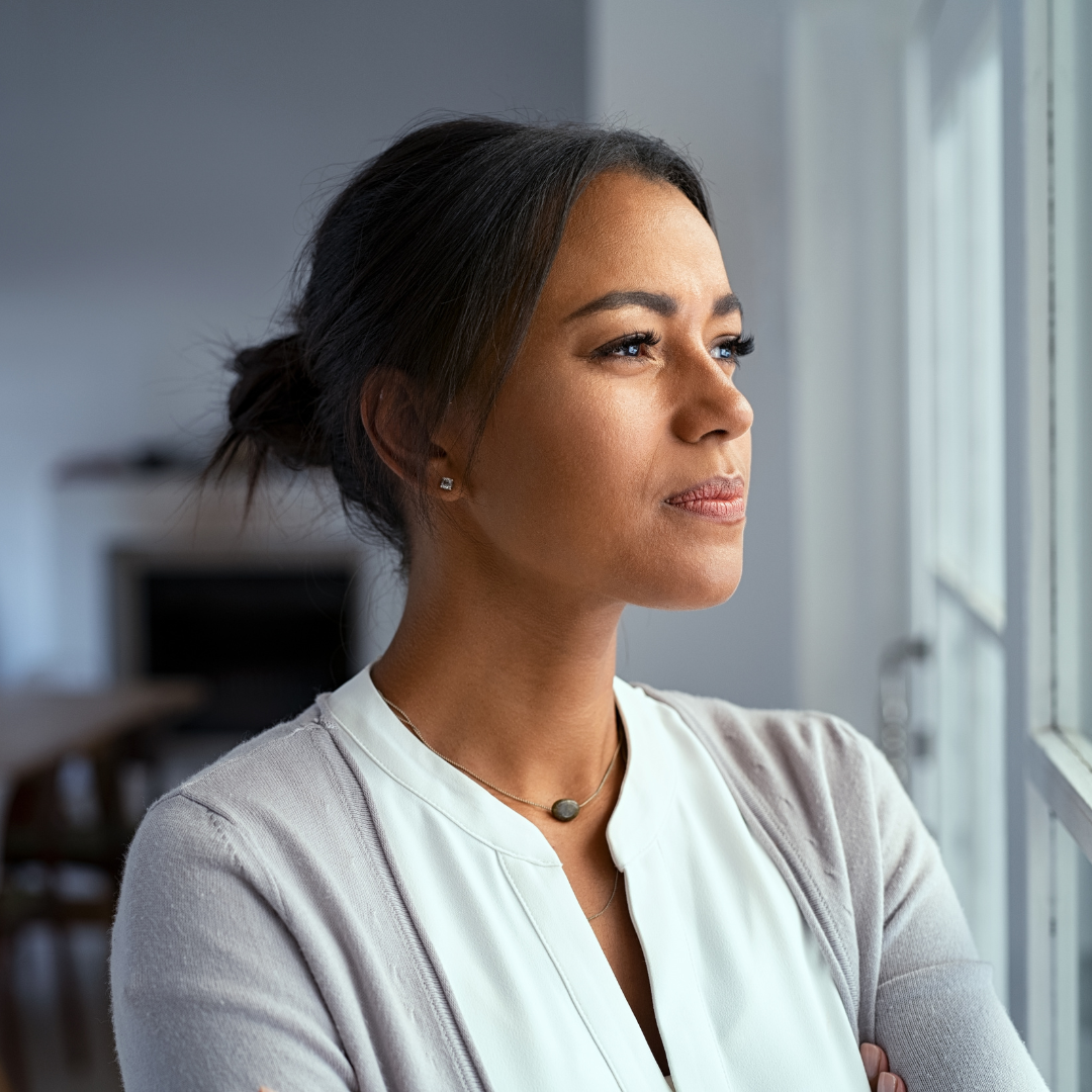 A woman with dark hair tied back, wearing a white top and a light gray cardigan, looking out the window with a contemplative expression.