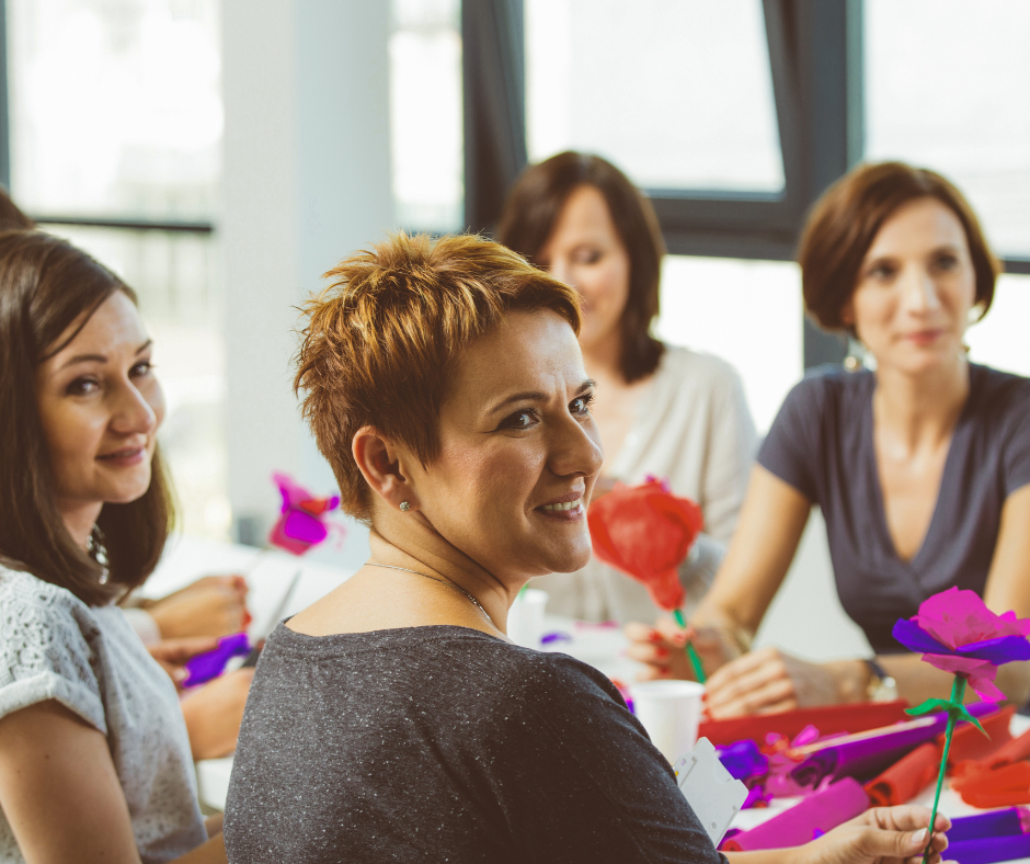 Group of five women sitting at a table decorated with colorful paper flowers, smiling and holding paper flowers in a bright room with large windows.