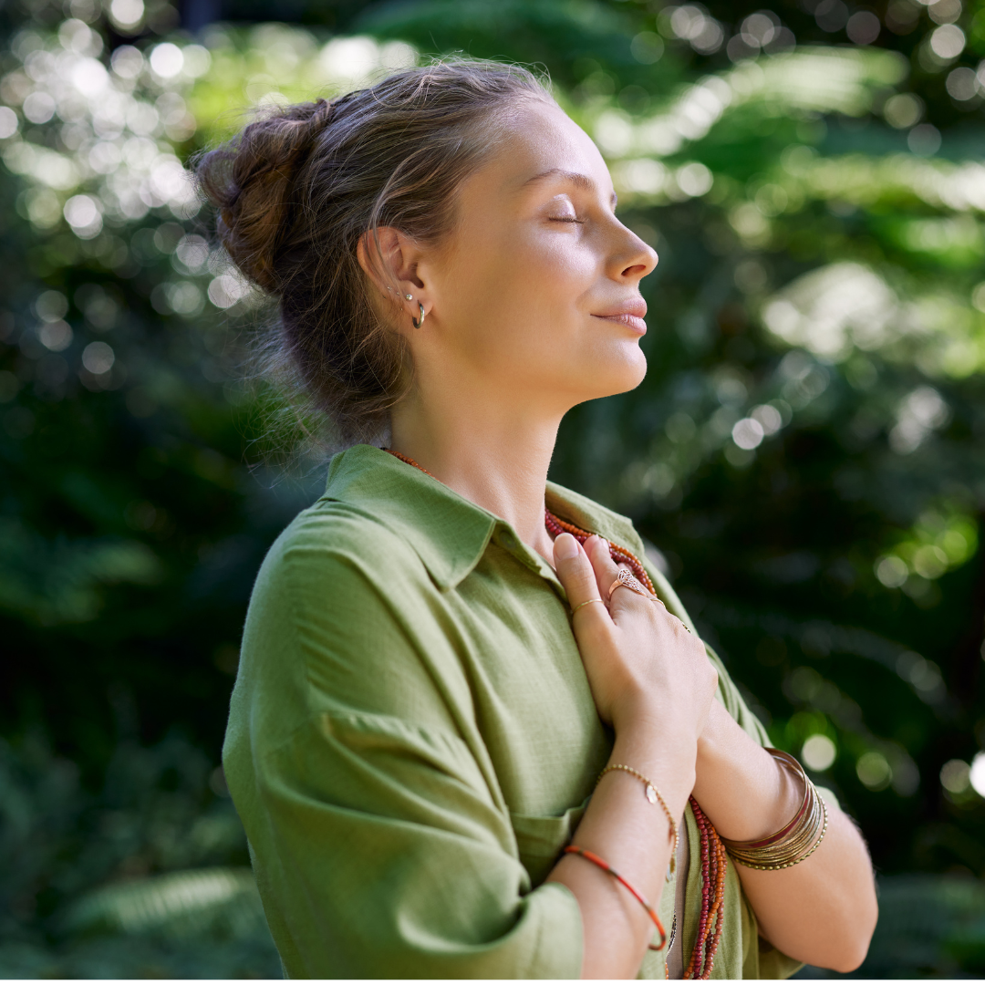 A woman with her eyes closed, smiling, with hands over her heart, standing outdoors surrounded by greenery.