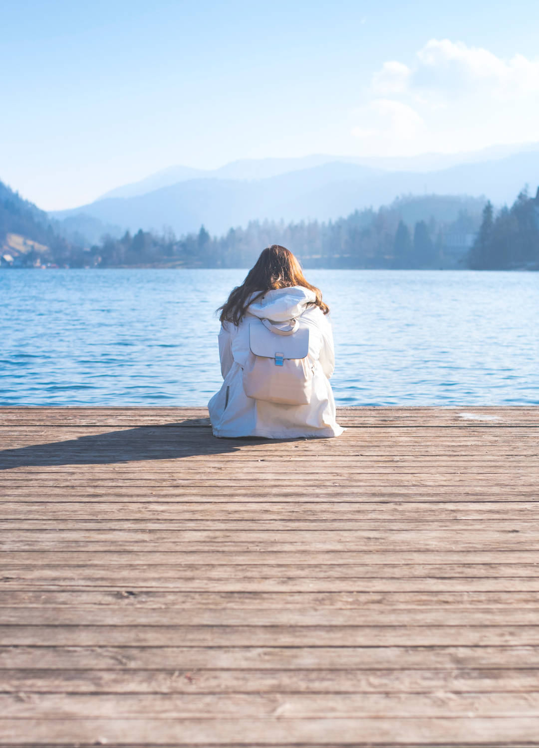 A person with long hair sitting on a wooden dock by a lake, facing away, with mountains in the background on a clear day.