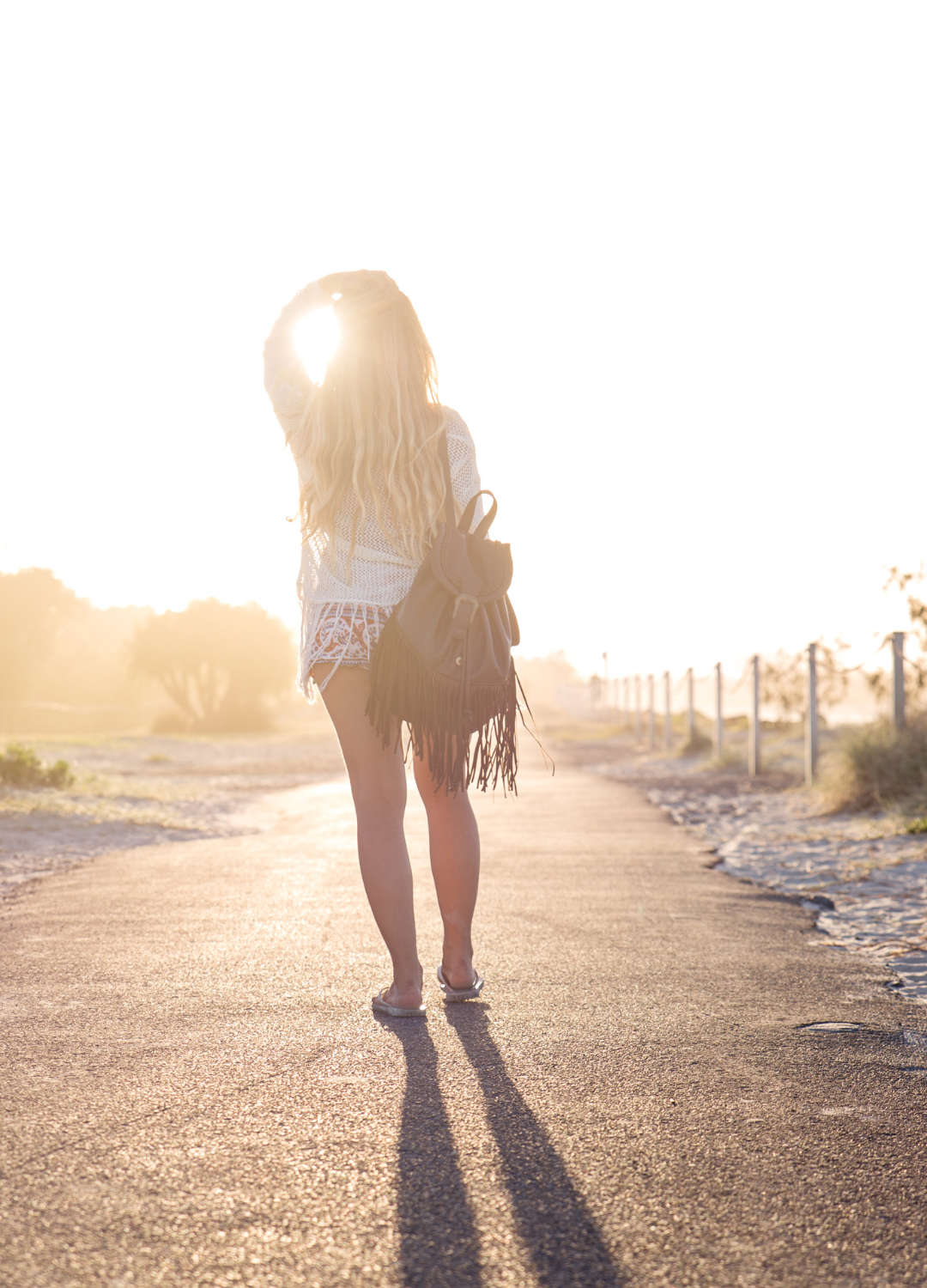 A woman with long blond hair walking on a dirt path during sunset, carrying a fringed backpack, wearing a white crochet top and shorts, with the sun shining directly behind her.