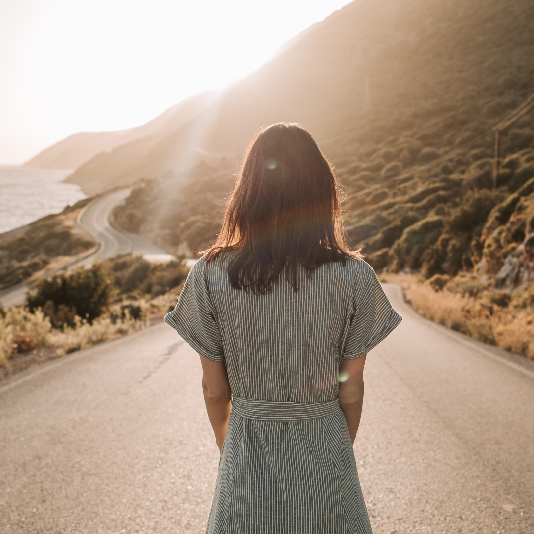A woman standing on a winding mountain road at sunset, facing away from the camera.